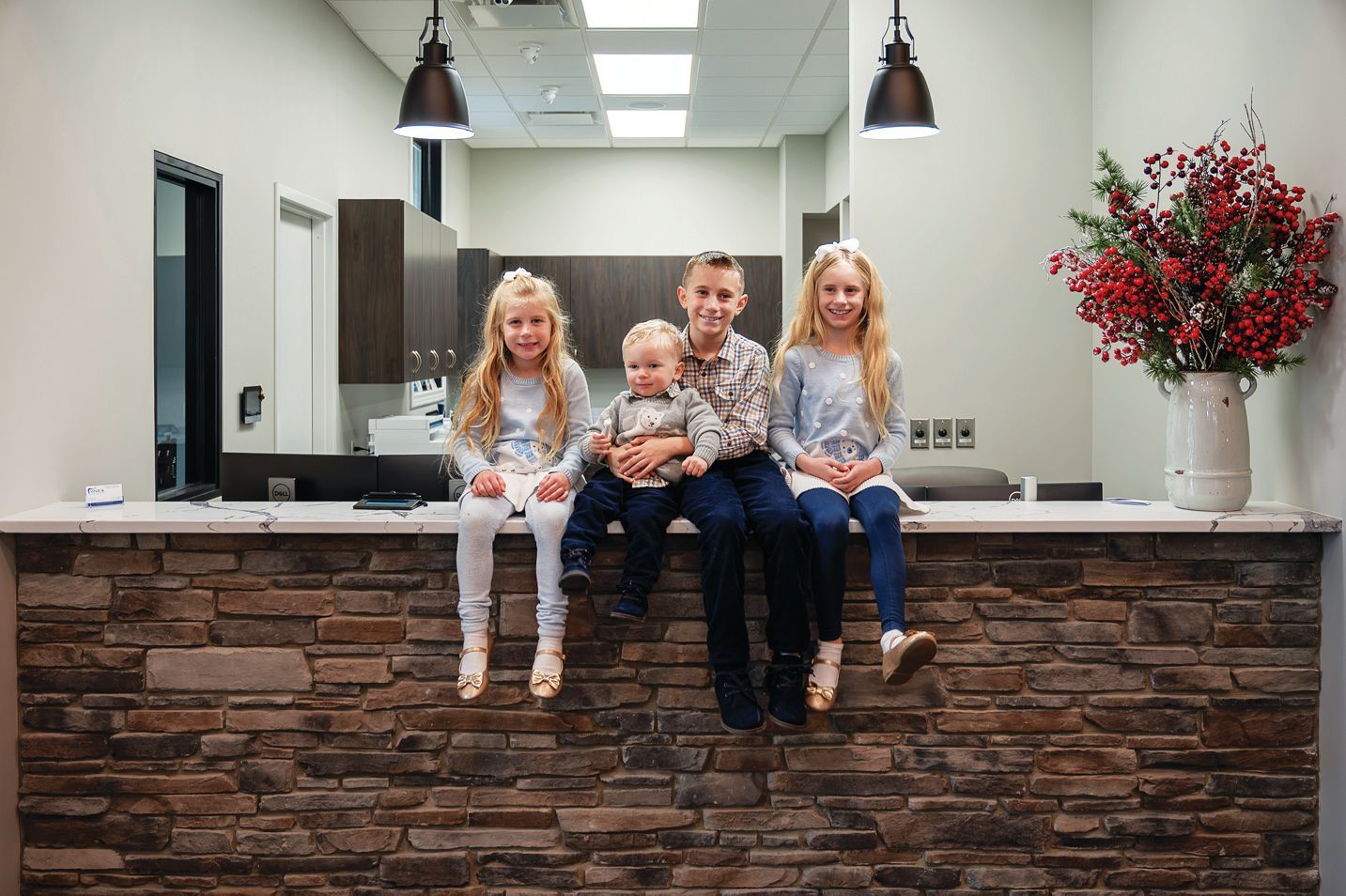 Four children sit on a stone counter in a modern, well-lit office lobby with a vase of red berries on the right.