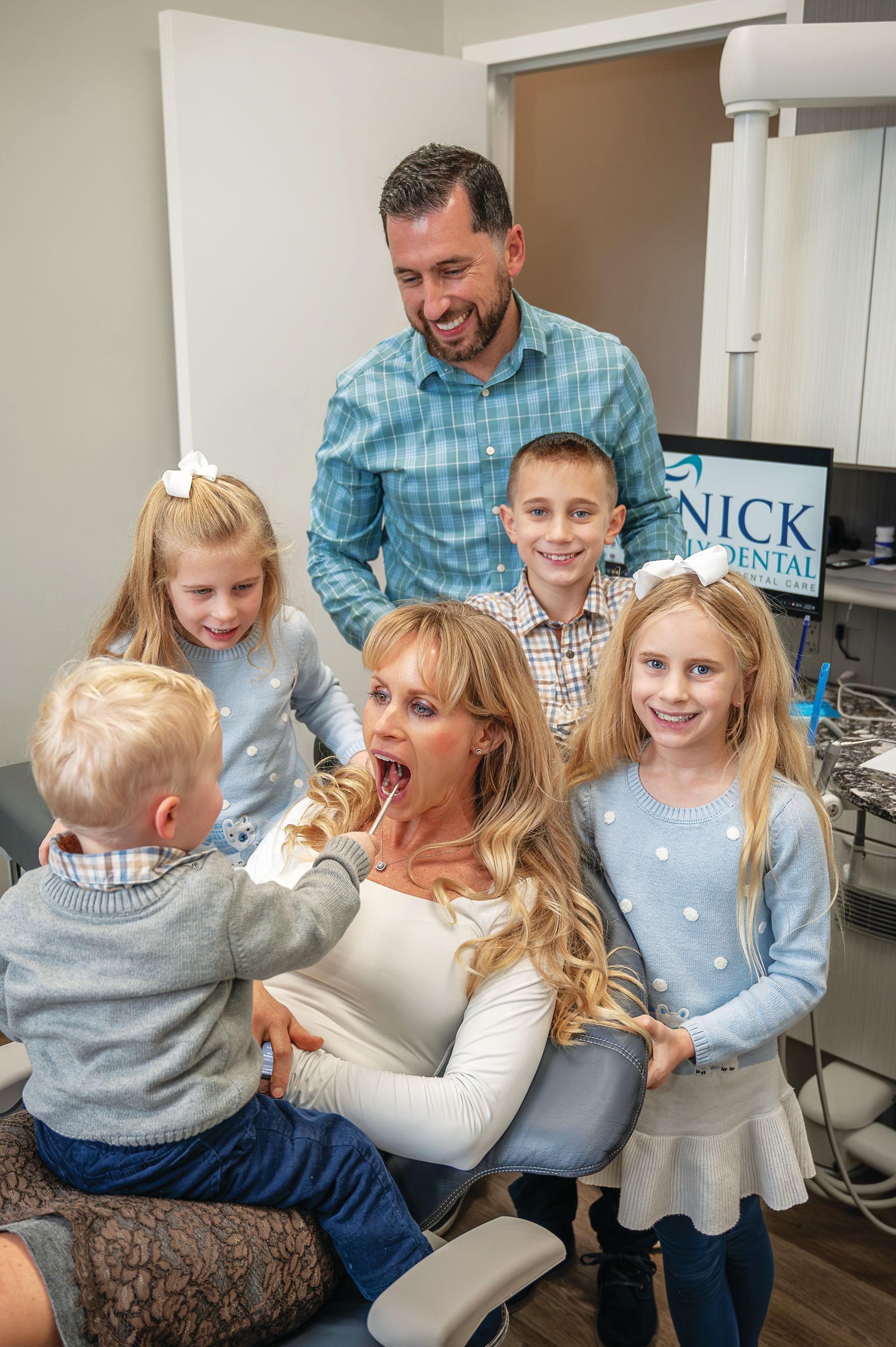 A family poses for a photo in a dental office as a young child playfully touches the open mouth of a person in the chair.