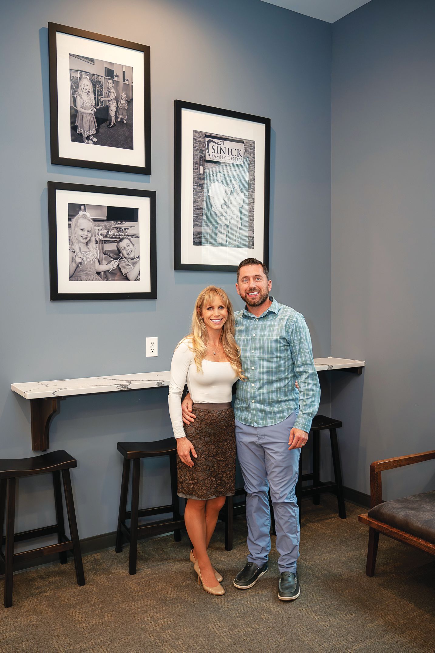 A smiling couple stands together indoors in front of a blue wall decorated with three framed black-and-white photos.