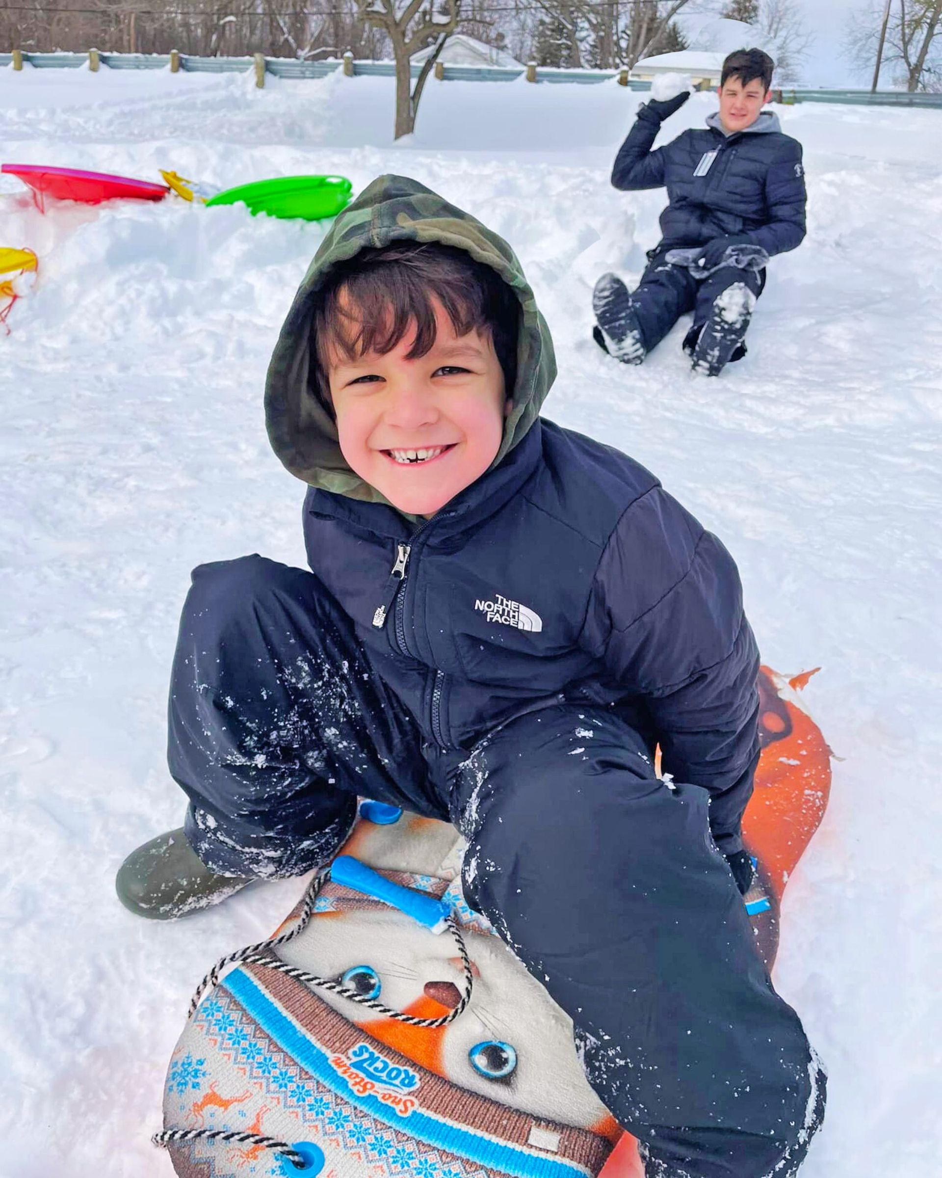 A smiling person in a camo hoodie sits on a snowman sled in the snow, with another person sitting in the background.