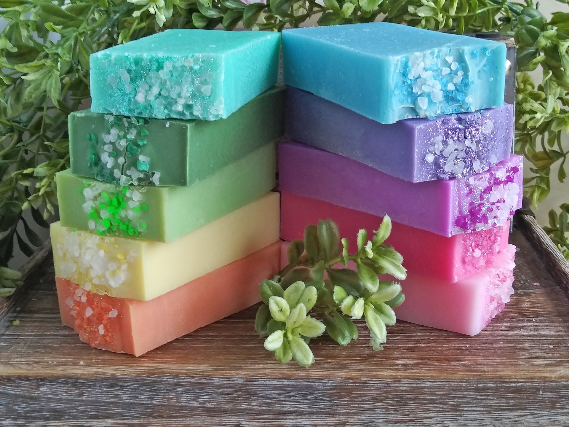 Two stacks of colorful, rectangular soap bars topped with decorative crystals, displayed on a wooden surface with greenery.