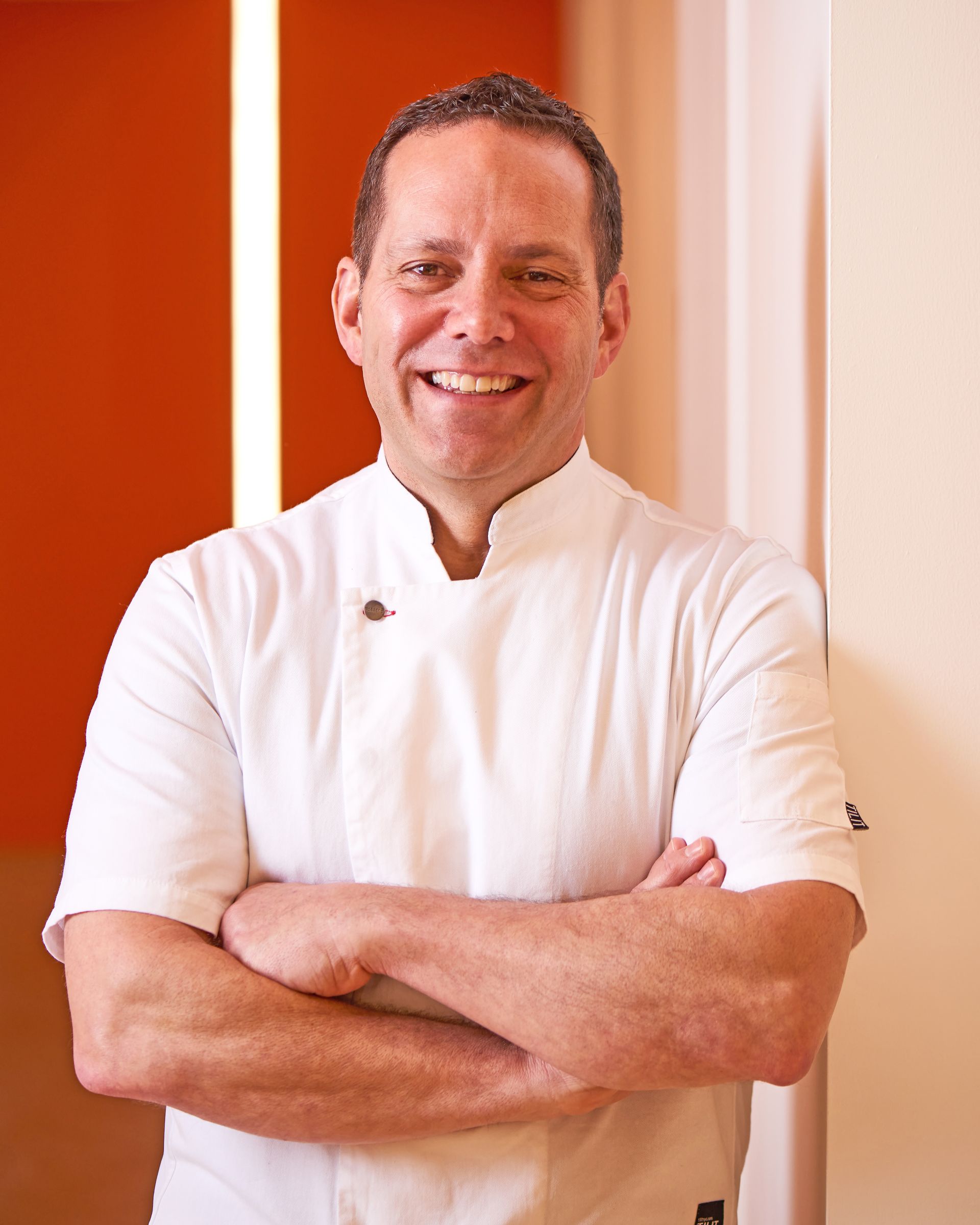 A smiling chef wearing a white uniform with arms crossed, set against a background of orange and white walls.