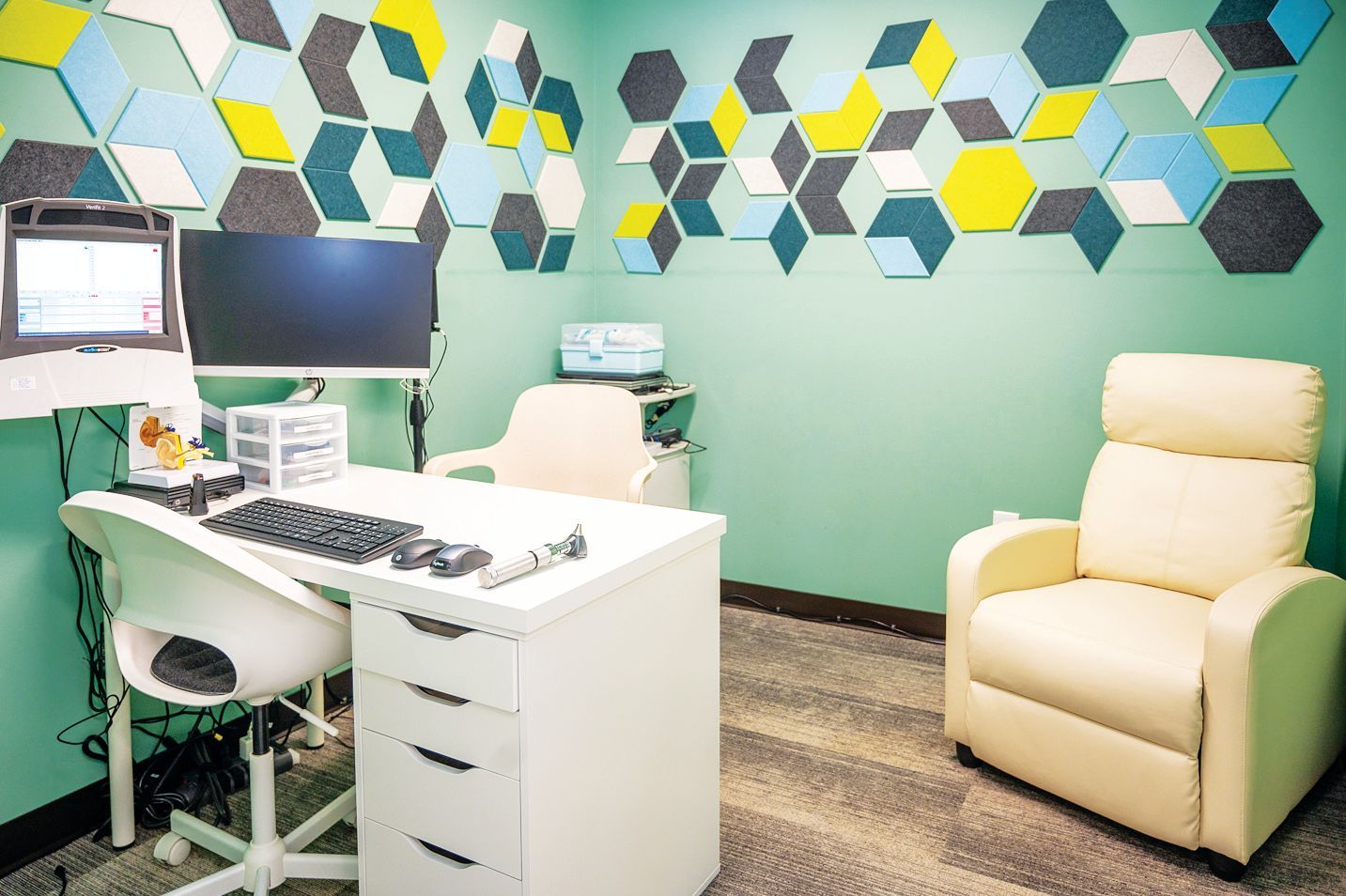 A medical office with a desk, computer, and chair, featuring green walls decorated with colorful geometric shapes.