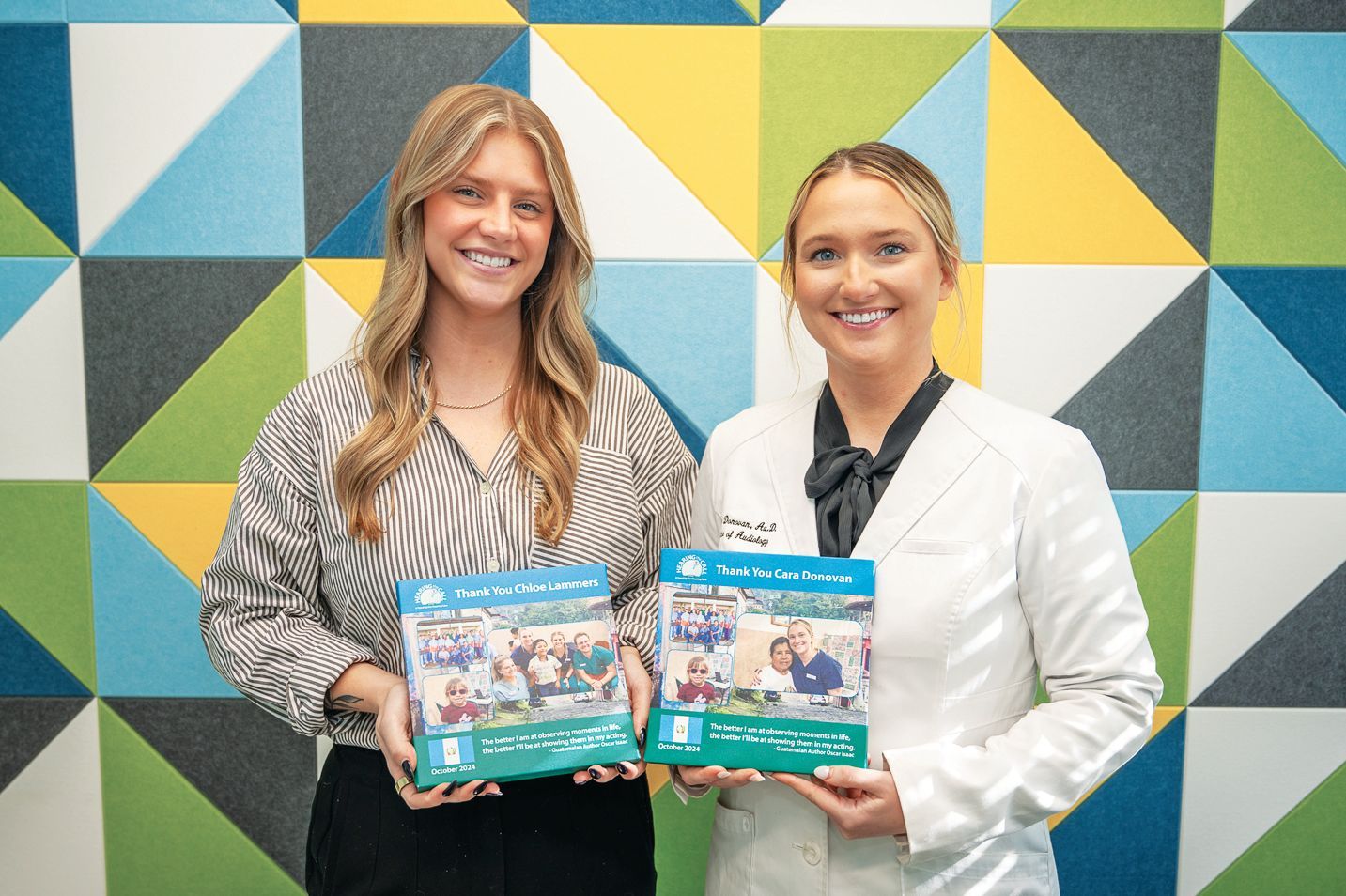Two smiling individuals stand against a multicolored geometric wall, each holding a blue informational brochure.