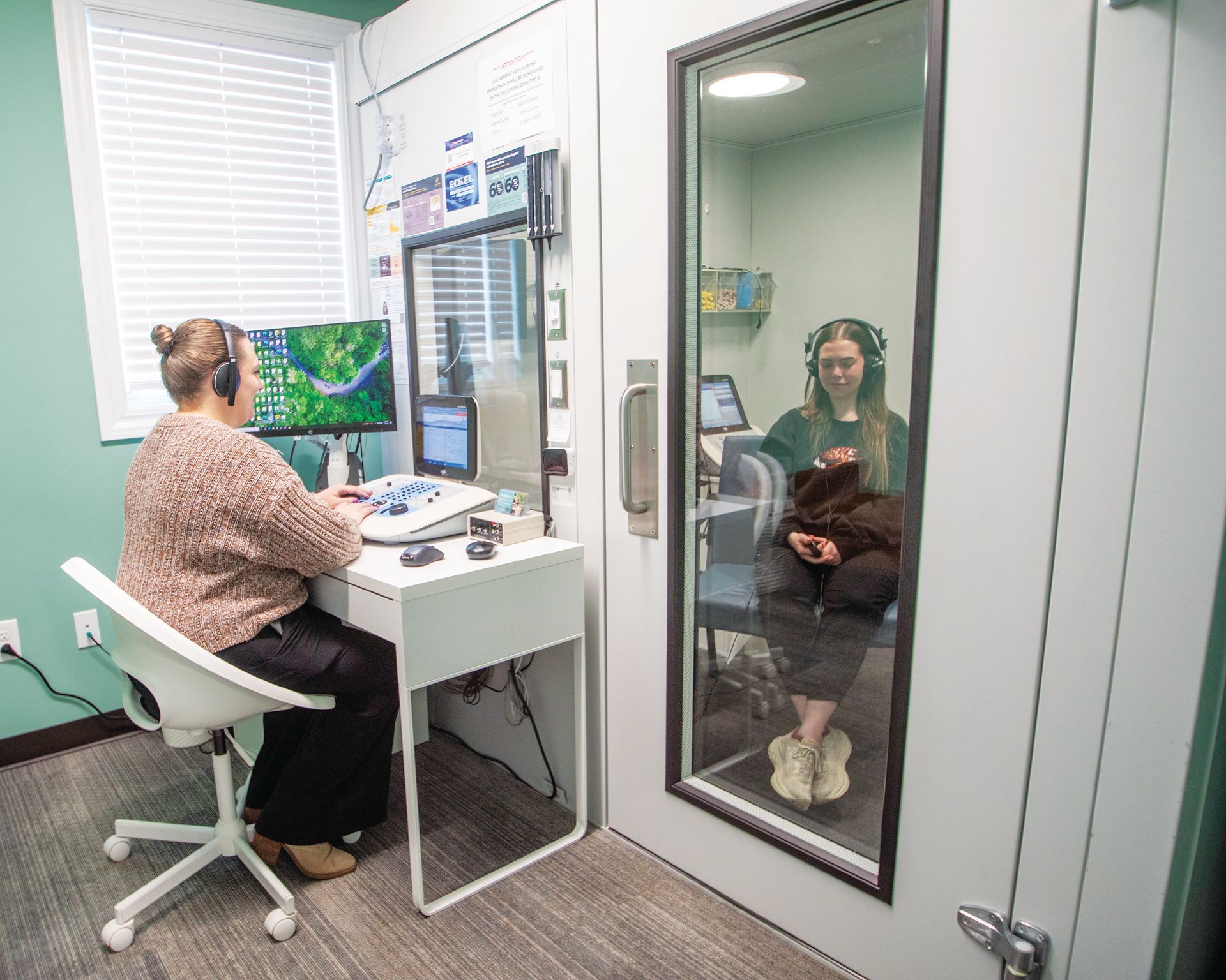 A clinician sits at a desk with a control panel and monitor, testing a patient wearing headphones in a soundproof booth.