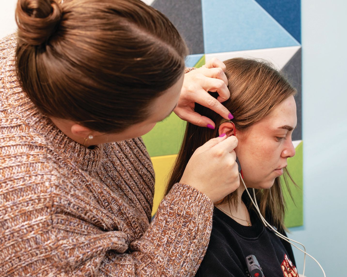 A person in a textured brown sweater inserts an in-ear hearing aid into another person's ear.