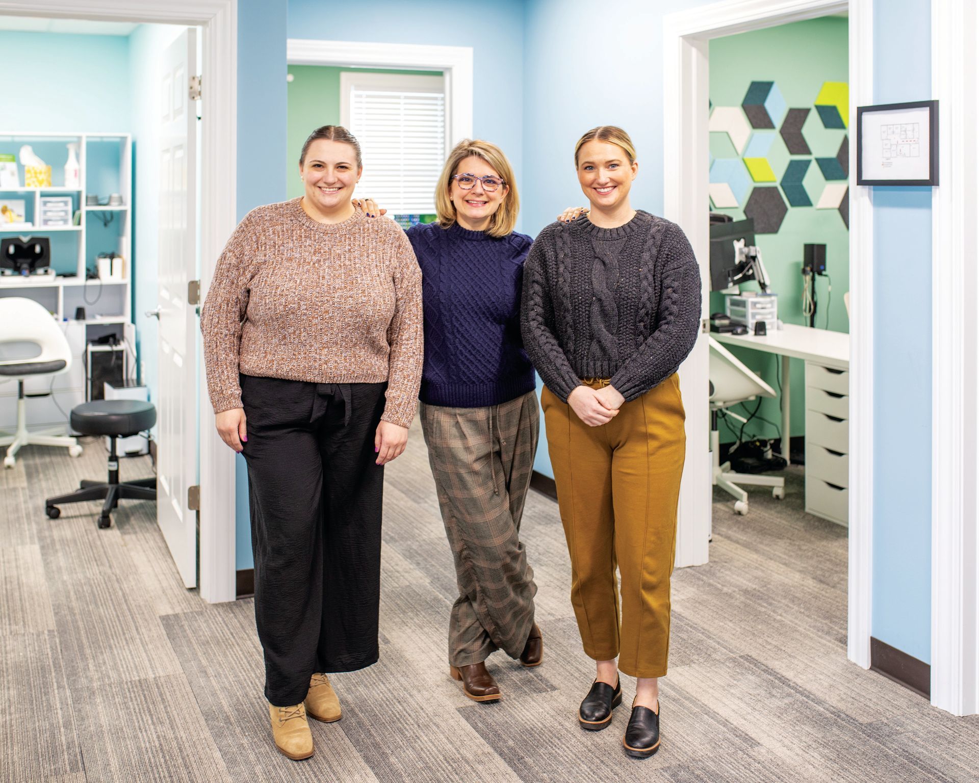 Three smiling people pose together in an office hallway wearing sweaters and casual trousers.
