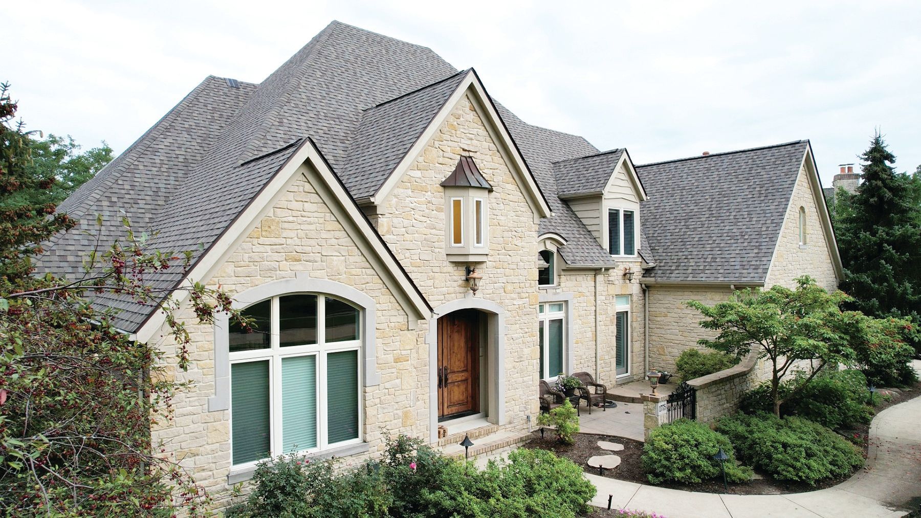 A two-story house with beige stone siding, a dark shingled roof, a wooden front door, and green landscaping.