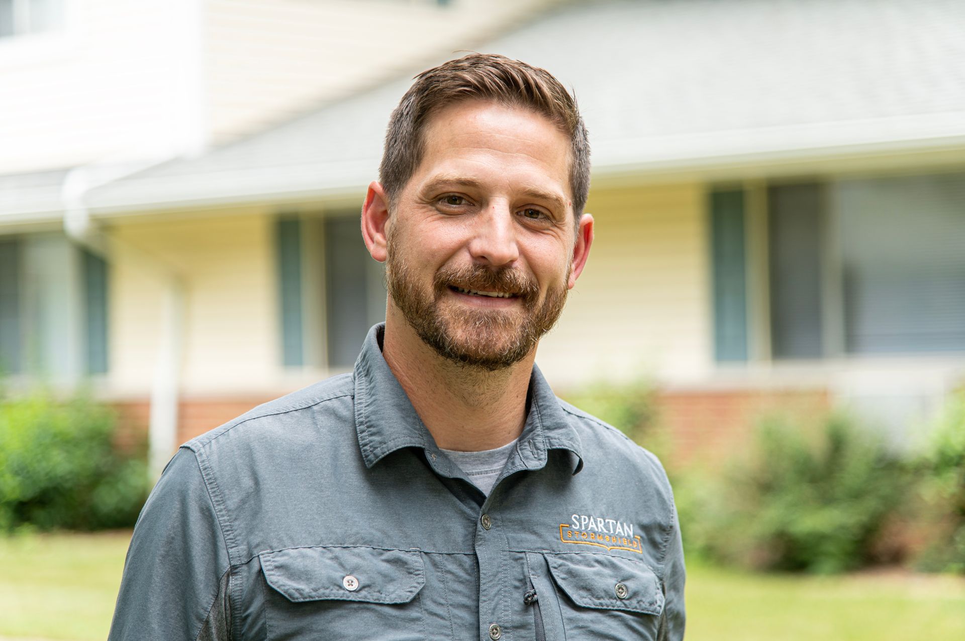 A person with a beard, wearing a gray button-down shirt, smiles outdoors in front of a blurry house and greenery.