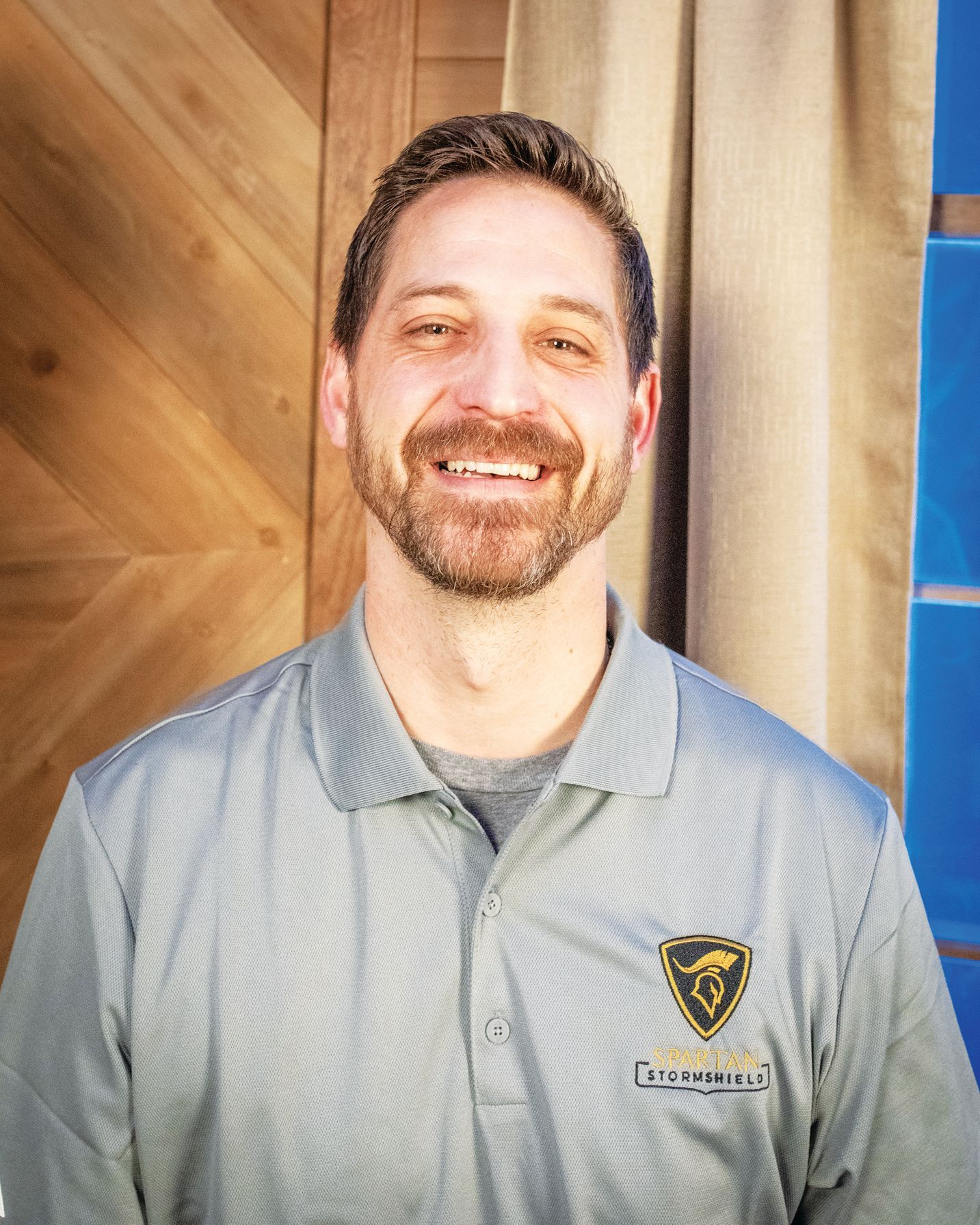 A person with a beard smiles while wearing a gray polo shirt with a black and gold kangaroo logo.