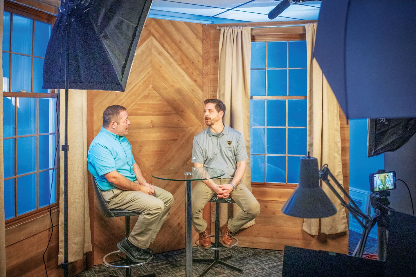 Two men sit on stools at a small table in a television studio, facing each other for a conversation with studio lights.