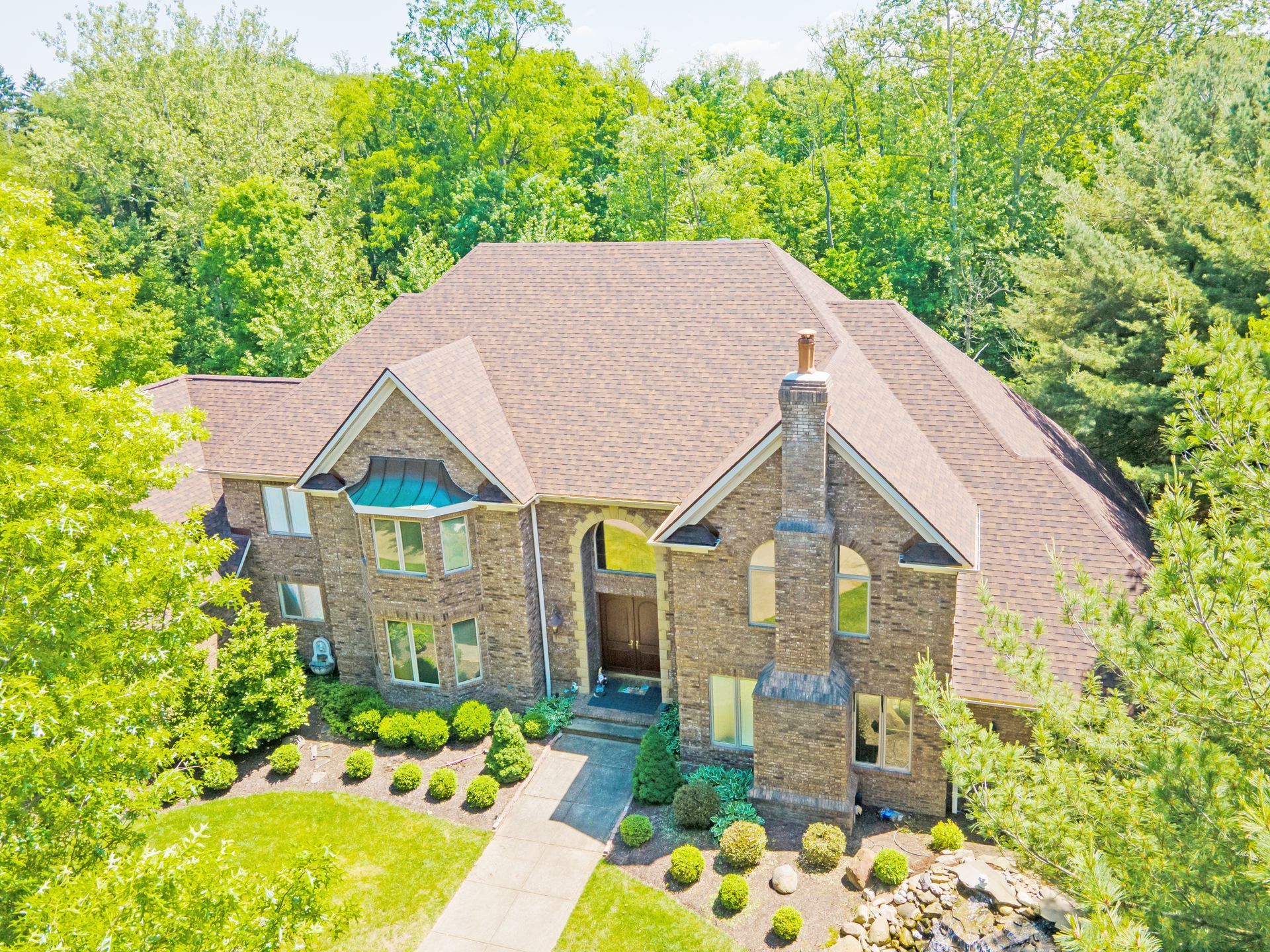 An aerial view of a large, two-story stone house with a brown shingled roof, surrounded by lush green trees and landscaping.