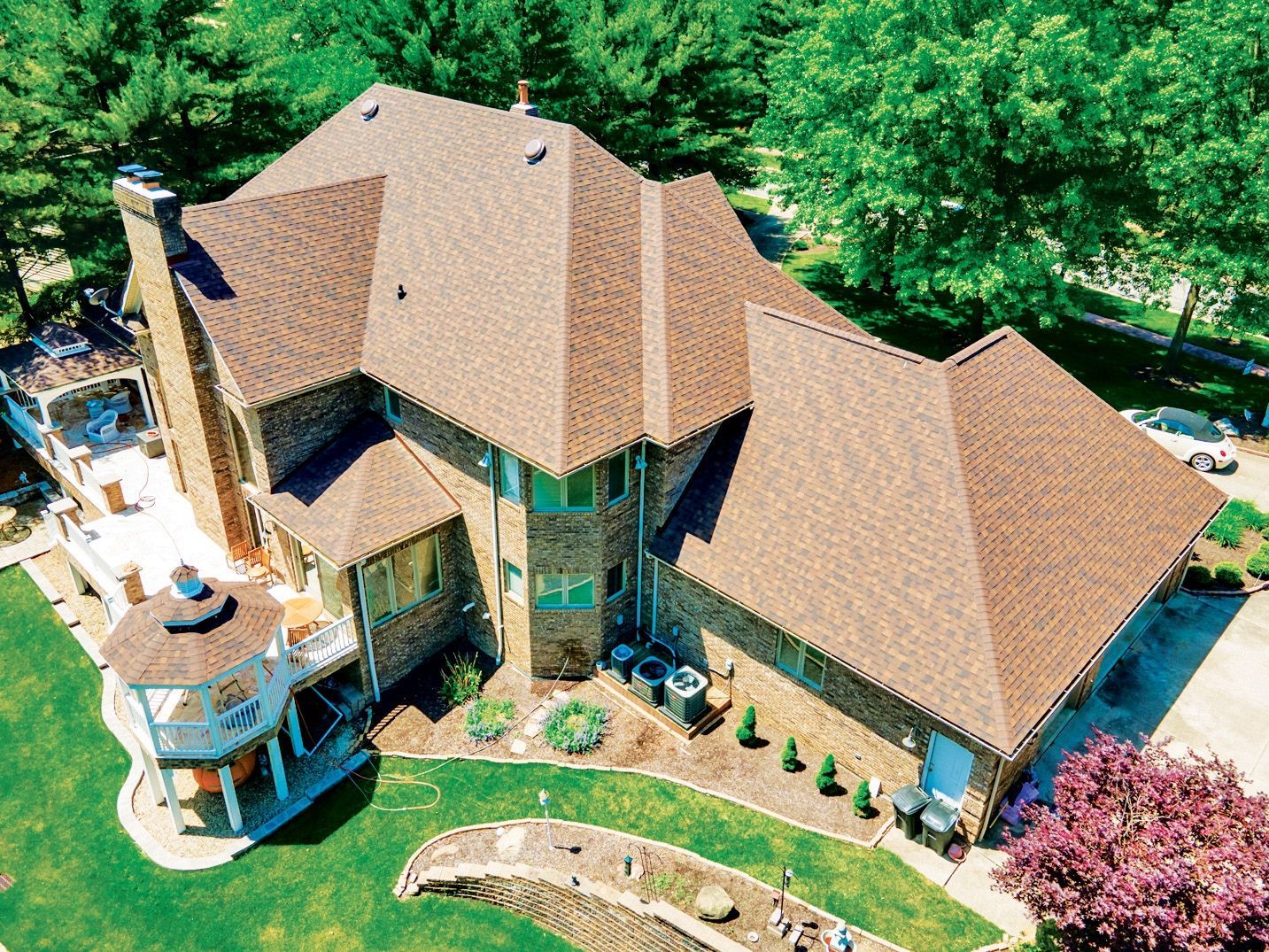 Aerial view of a large, brown-roofed brick house with a gazebo and a patio, surrounded by trees and a lawn.
