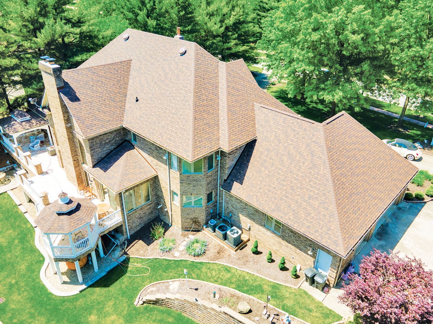 An aerial view of a stone house with a complex brown shingled roof, a gazebo, and a landscaped yard with trees.