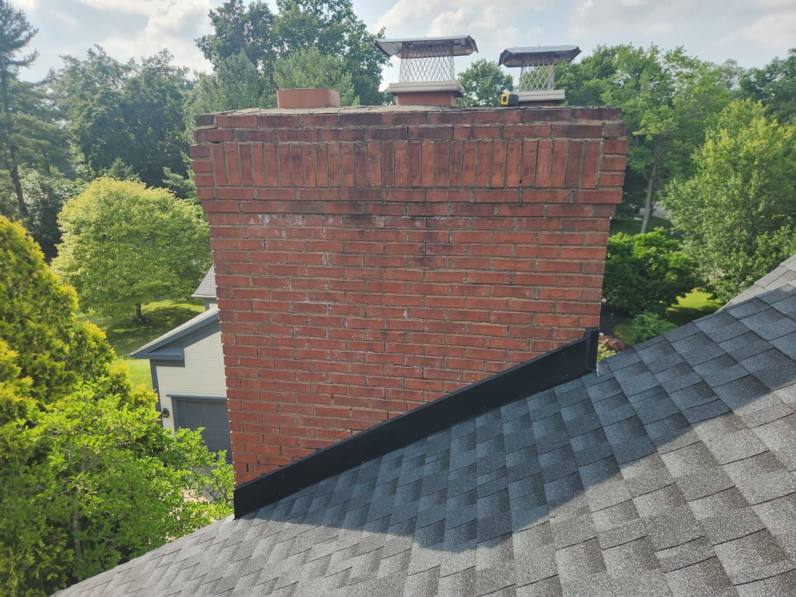 A brick chimney on a shingled roof with metal flashing at the base and two chimney caps on top.