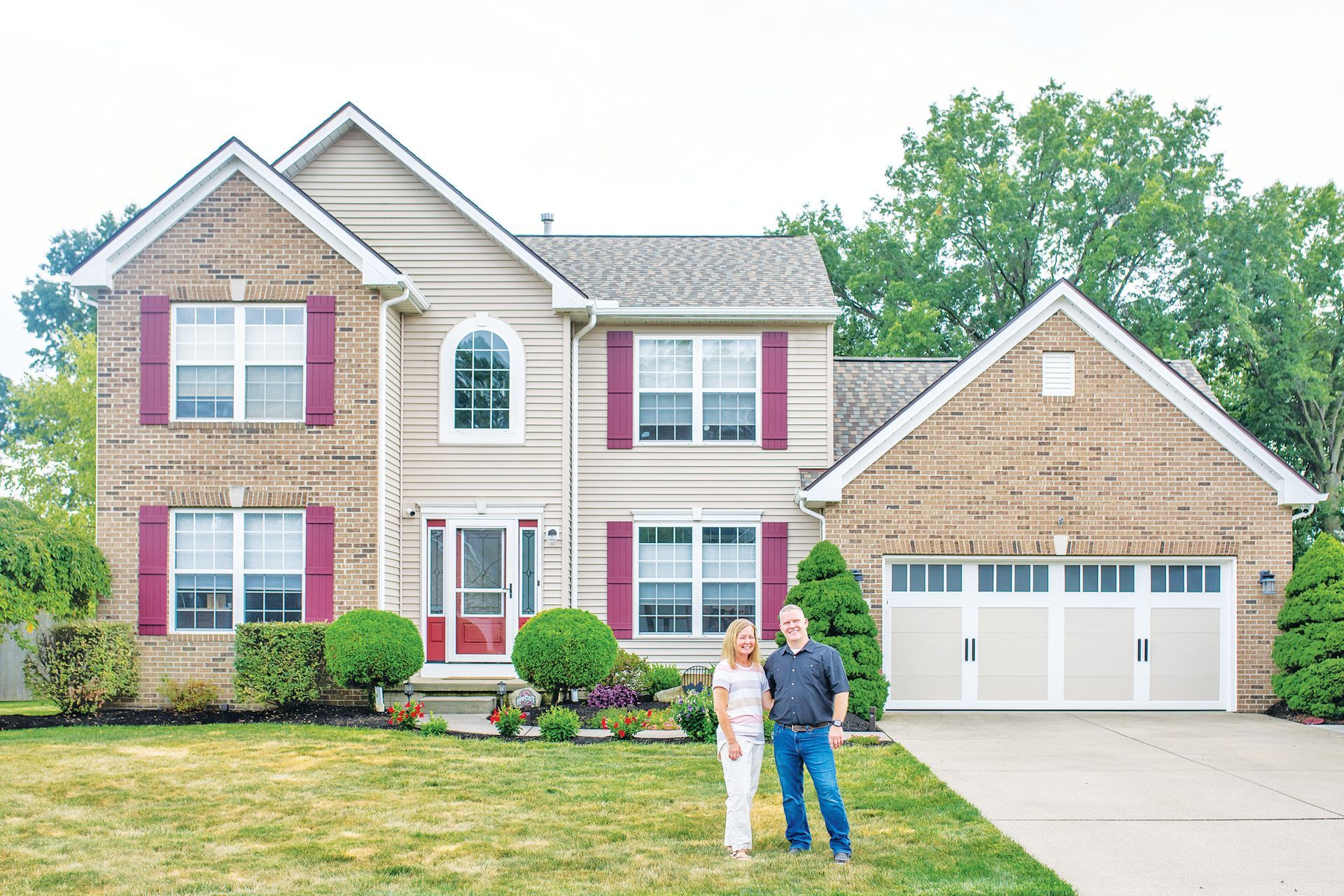 A couple stands in the front yard of a two-story beige brick and siding house with red shutters and a three-car garage.