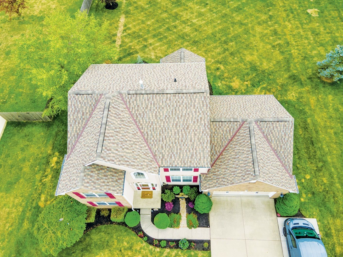 Aerial view of a residential house with a beige shingled roof, red shutters, and a concrete driveway in a green yard.