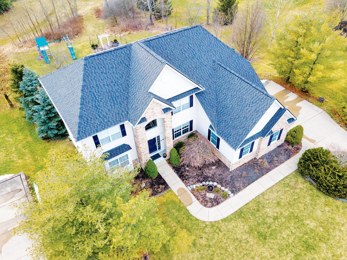 An aerial view of a two-story house with a blue shingled roof, light brick exterior, and a surrounding yard with trees.