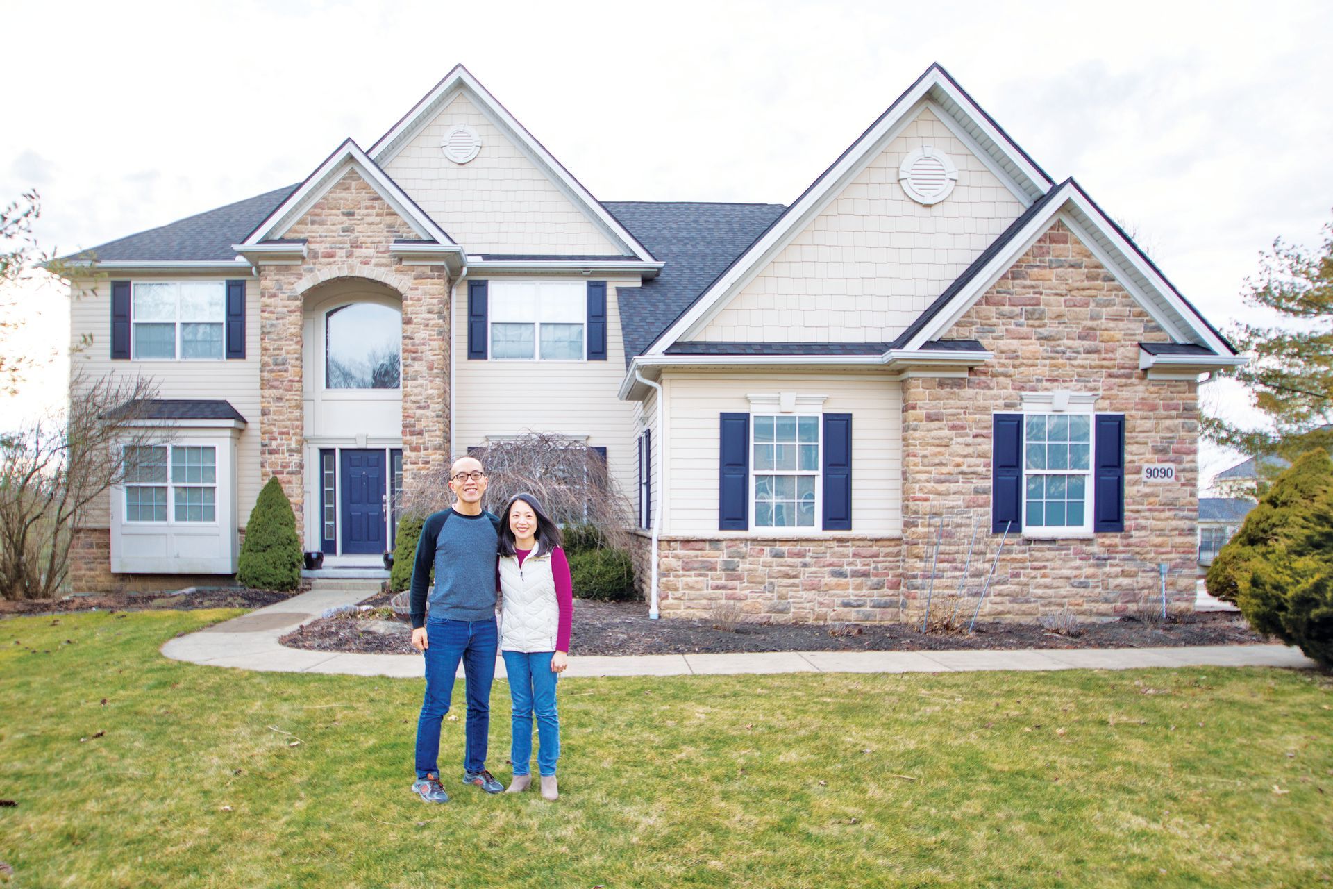A couple stands together on a lawn in front of their two-story suburban home with stone and light-colored siding.