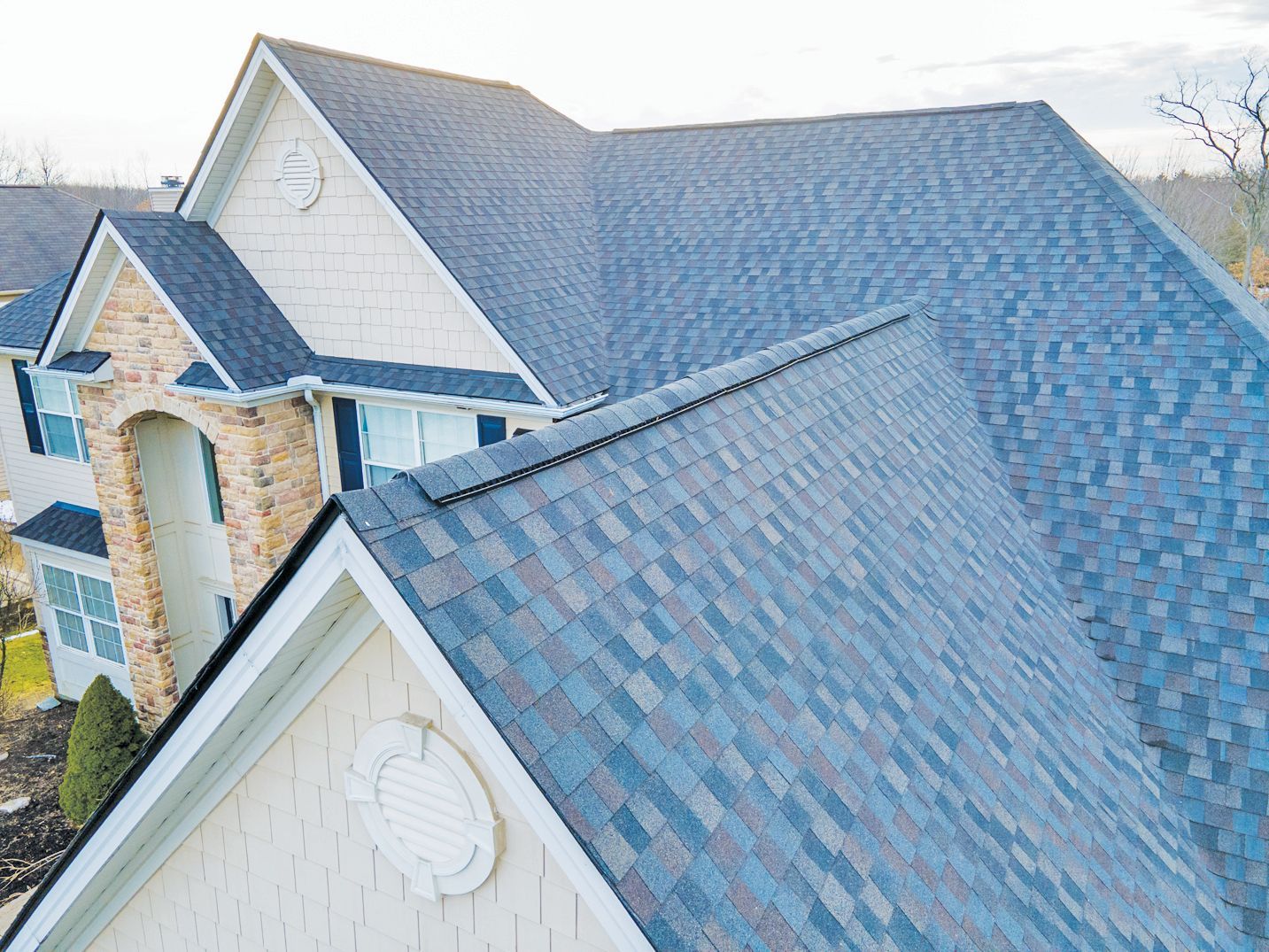 An aerial view of a multi-gabled house with a textured blue-grey shingled roof and cream-colored exterior walls.