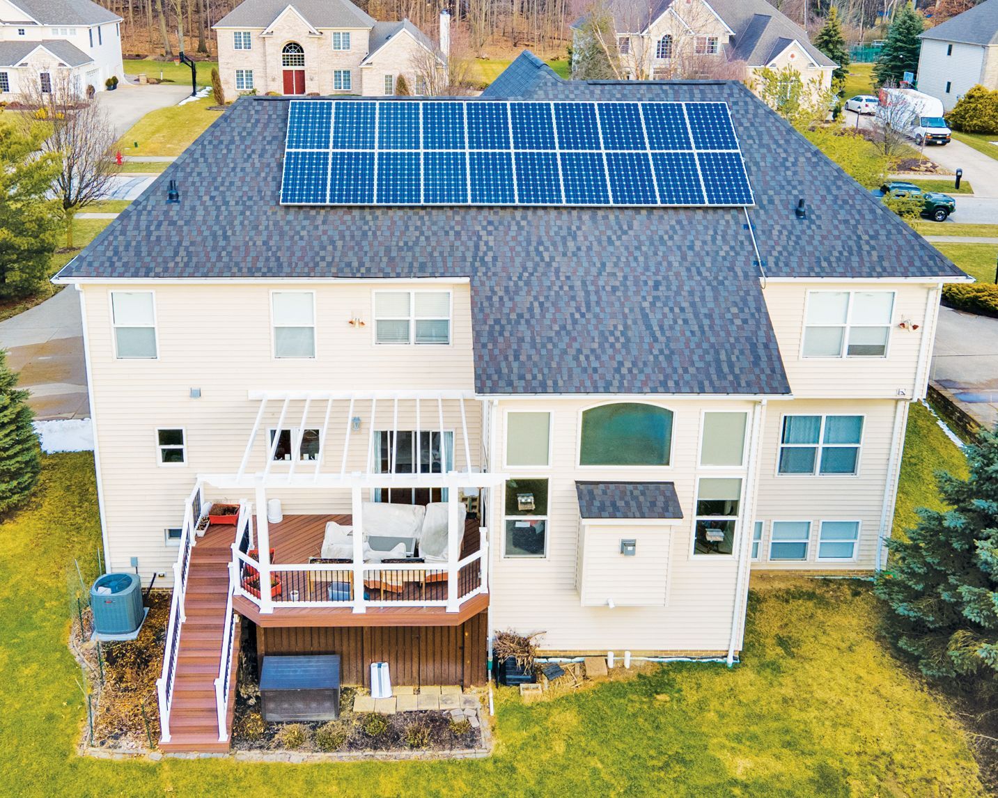 A two-story tan house with a rooftop solar panel array, a white pergola-covered deck, and a wooden staircase.