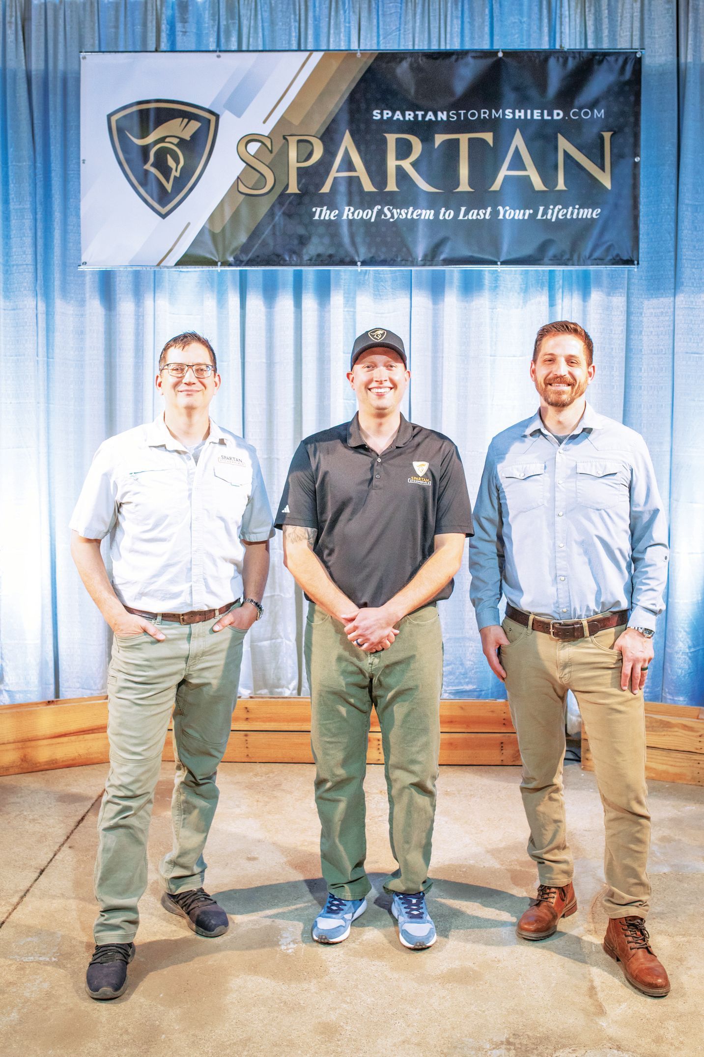 Three men stand side-by-side in front of a blue Spartan-branded banner at an event.