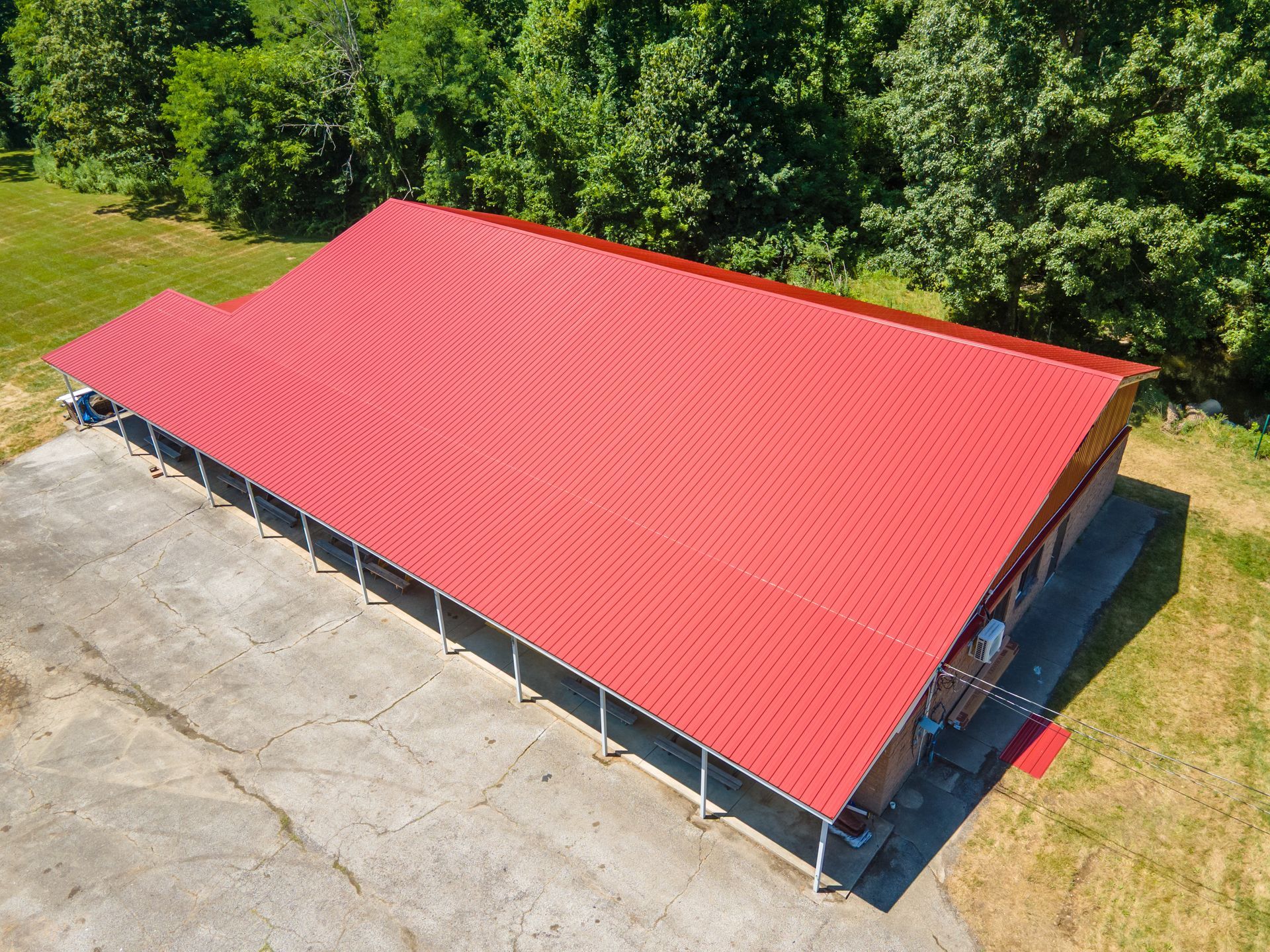 An aerial view of a long rectangular building with a bright red metal roof, situated next to a paved lot and green trees.