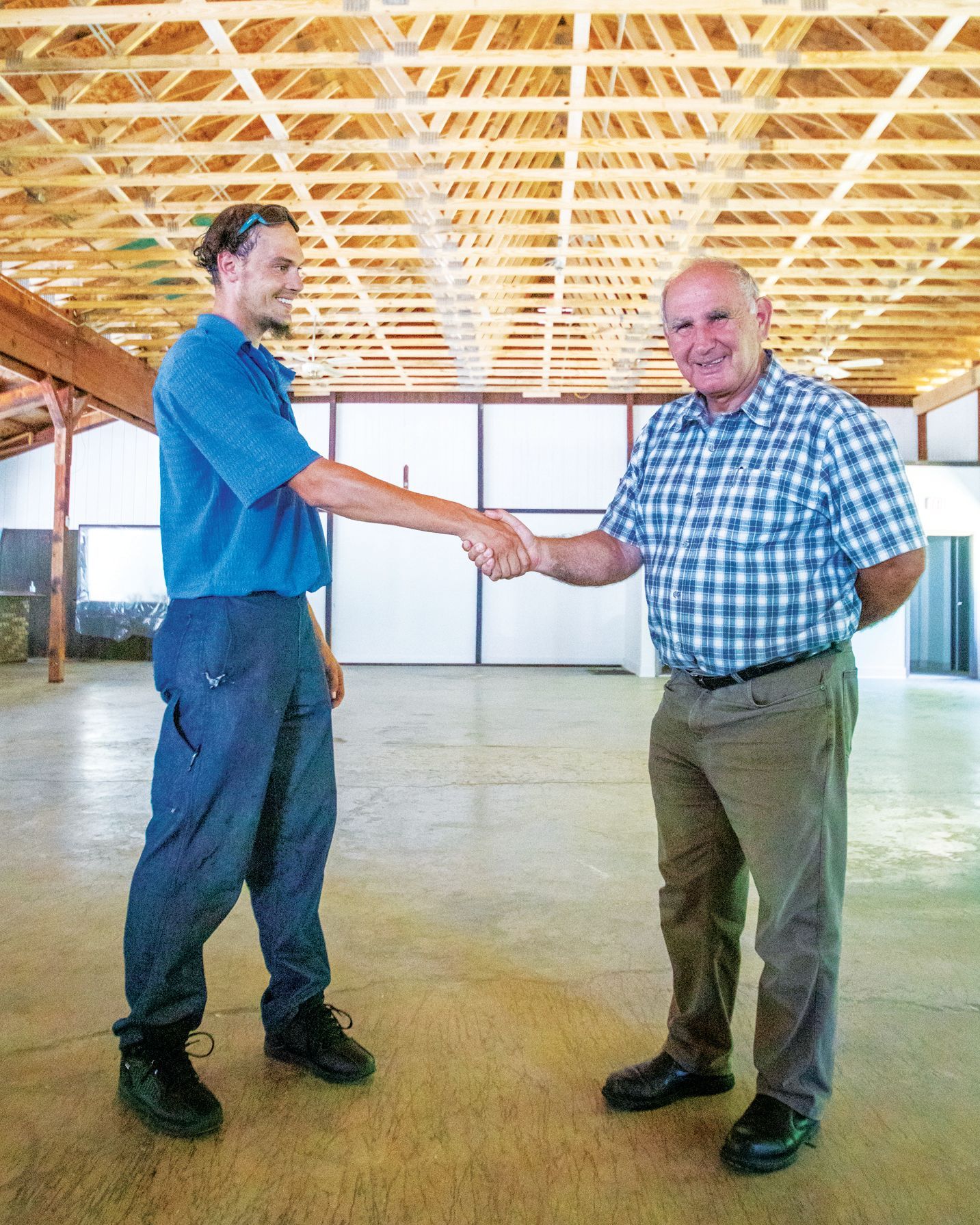 Two people shake hands in the middle of a large, empty, industrial-style building with a wooden roof structure.
