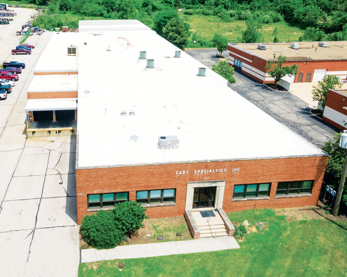 Aerial view of a red-brick commercial building with a white roof, surrounding parking, and nearby greenery.