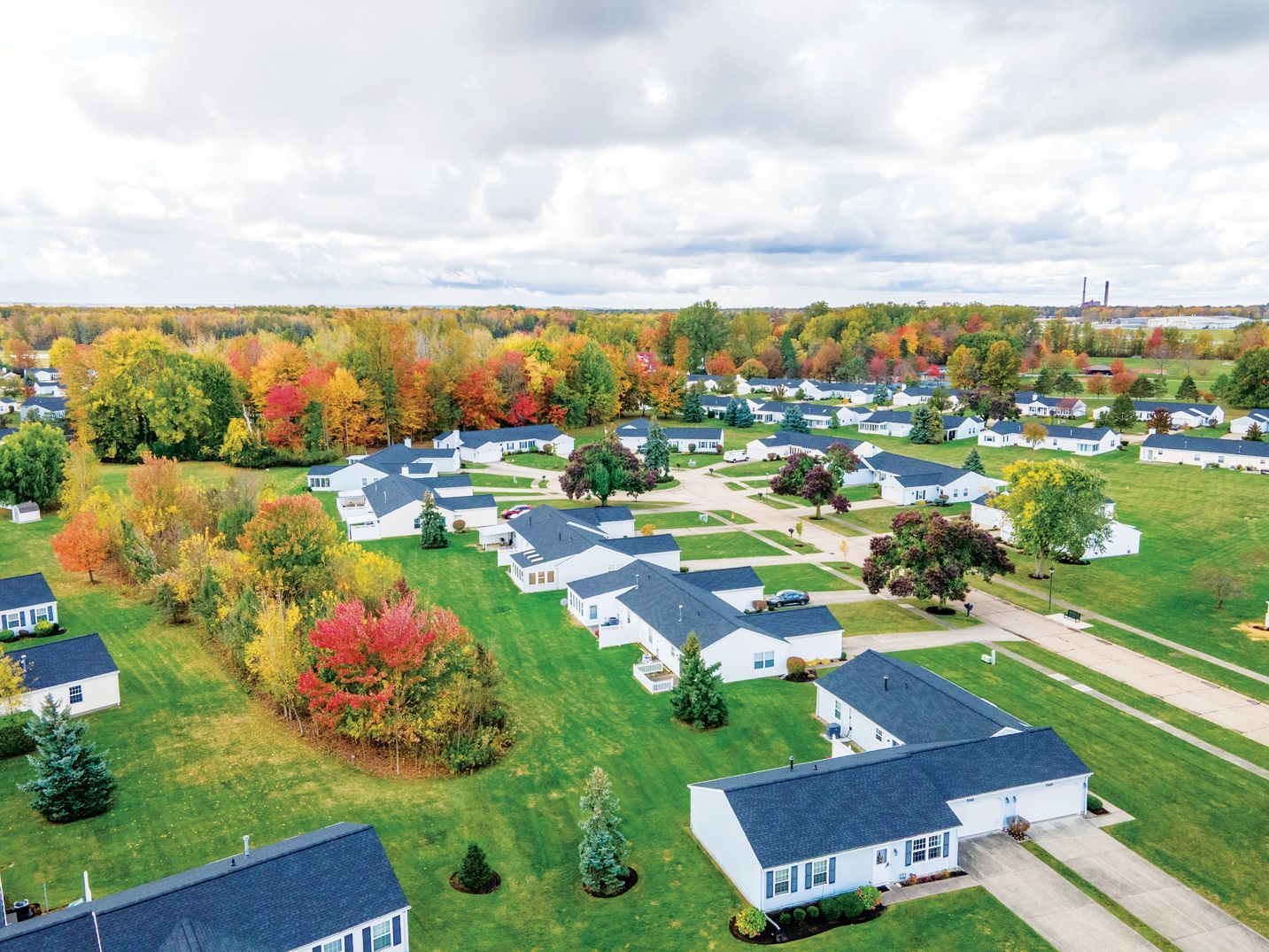 Aerial view of a residential neighborhood with white houses, dark roofs, and fall trees set against a cloudy sky.