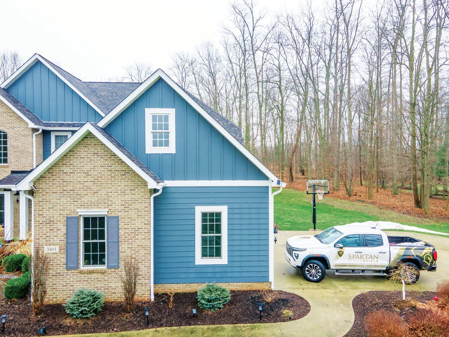 A blue-sided home with stone accents and a white utility truck parked in the driveway near a basketball hoop and trees.
