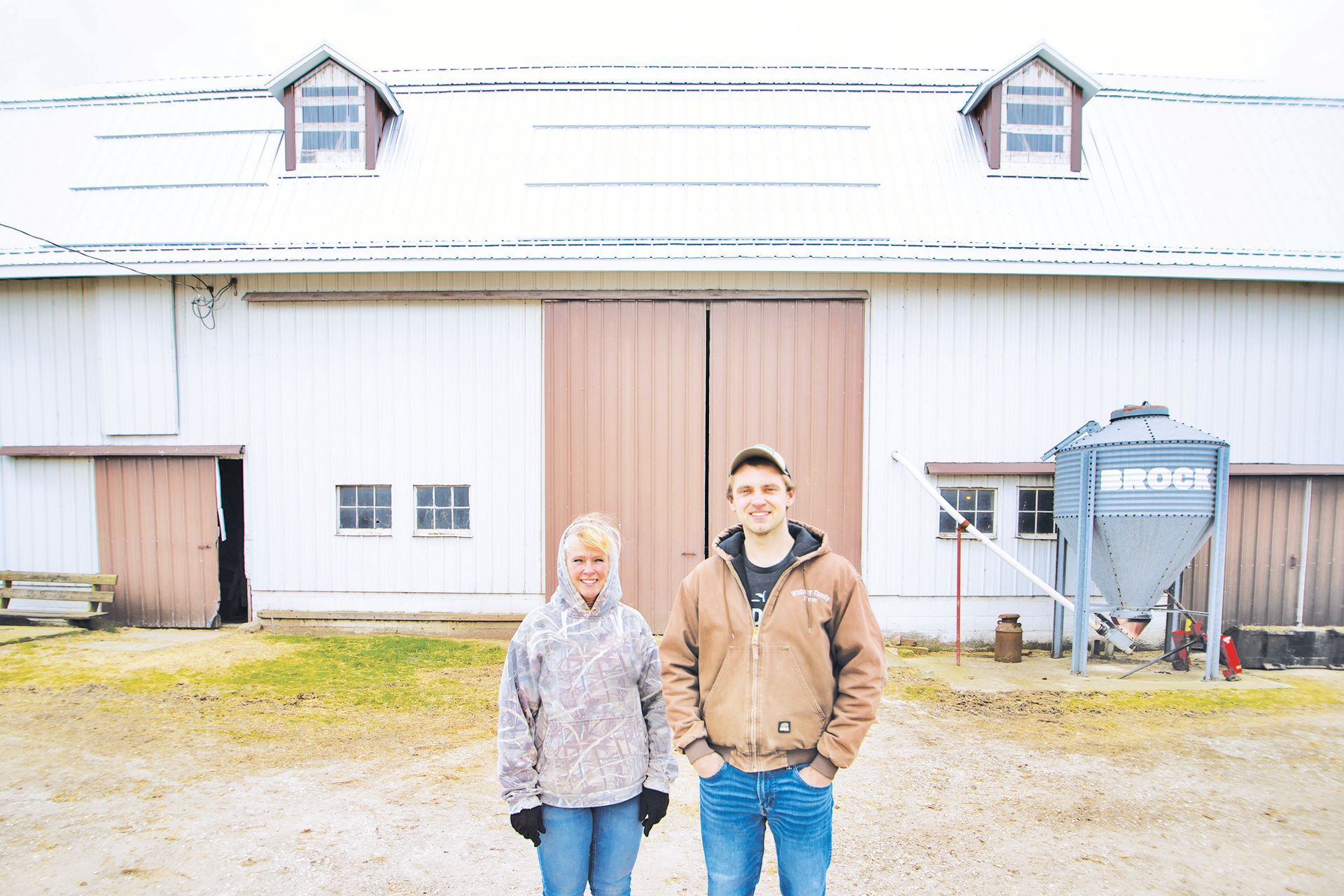 Two people stand smiling in front of a white barn with a snowy roof, a brown sliding door, and a metal silo.