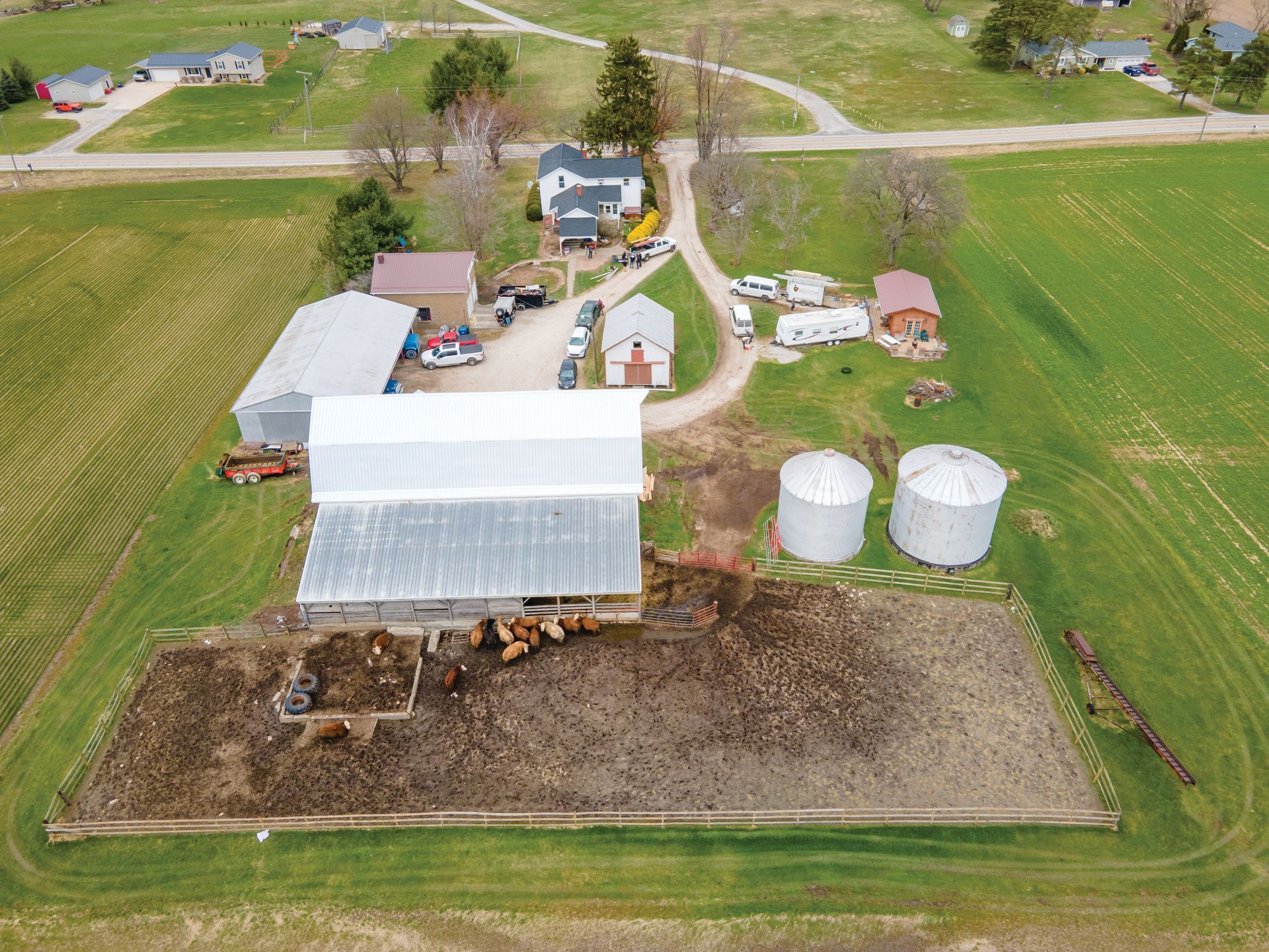 Aerial view of a farm featuring a large white barn, cattle pens, two grain silos, and several outbuildings on green land.