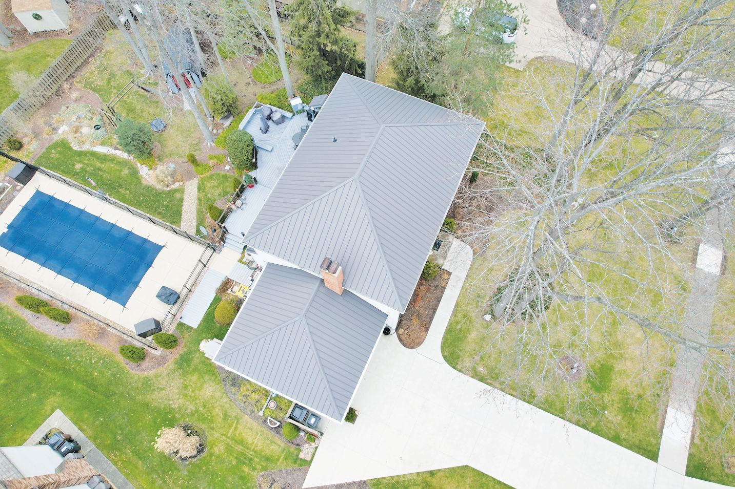 An aerial view of a house with a gray roof, an adjacent pool with a blue cover, a paved driveway, and surrounding lawn.