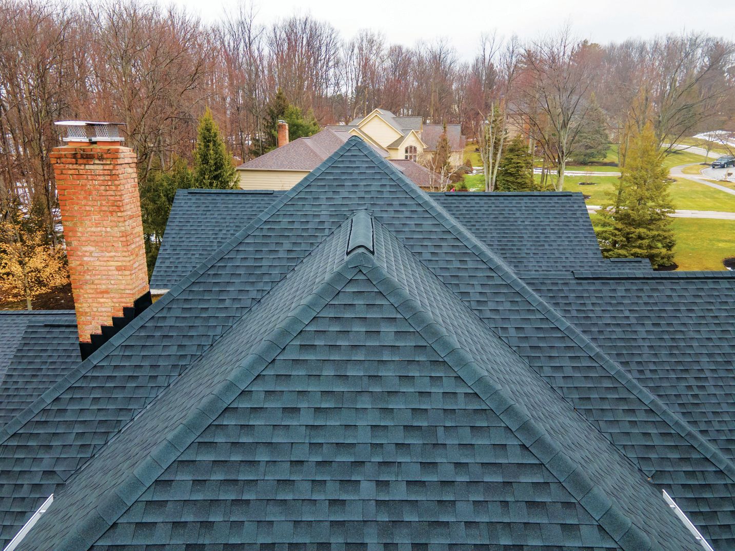 High-angle view of a dark shingled roof with a brick chimney and multiple gables, set against a background of trees.