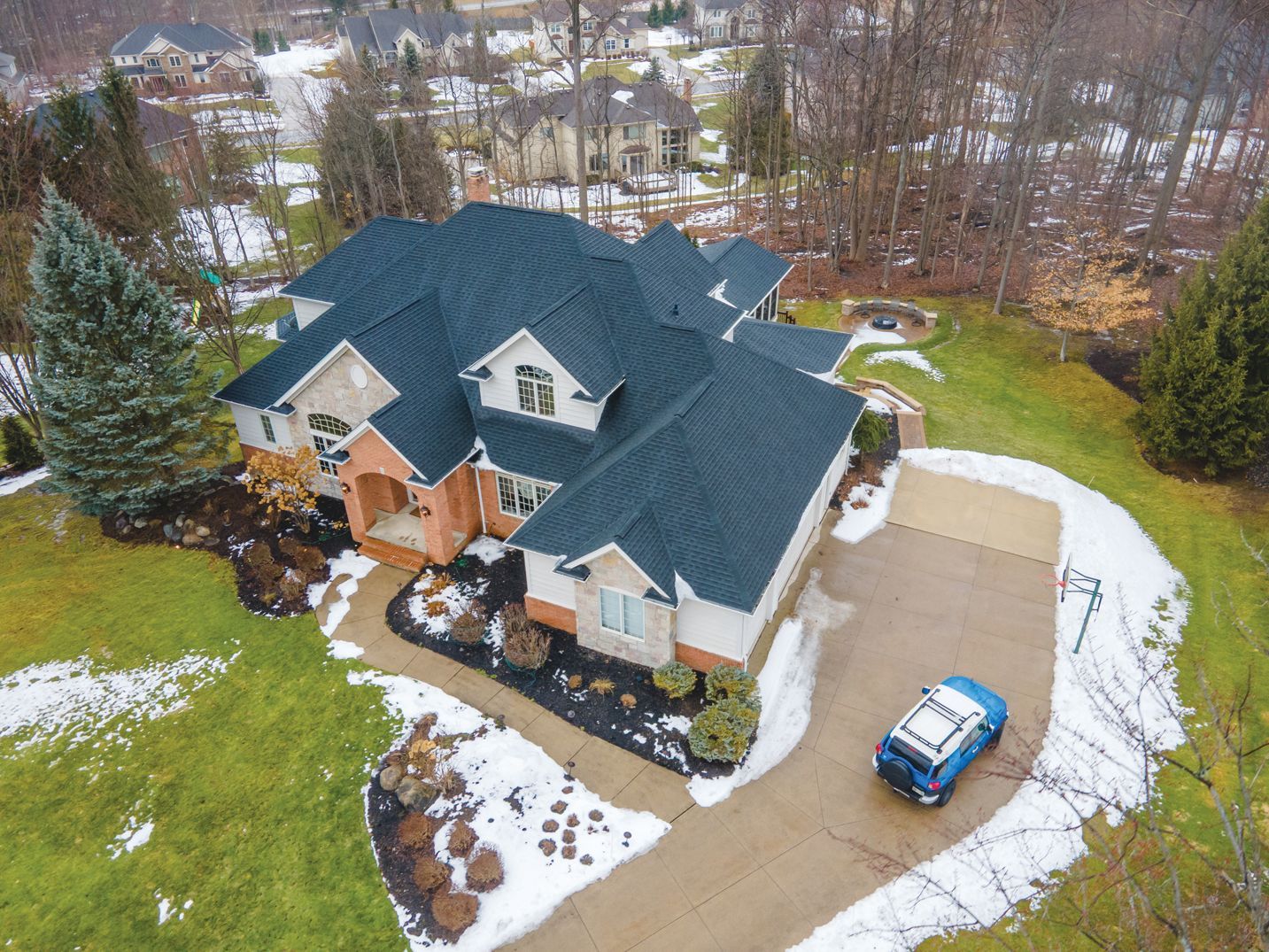 An aerial view of a large suburban house with a dark roof, surrounded by a lawn with patches of snow and a parked car.