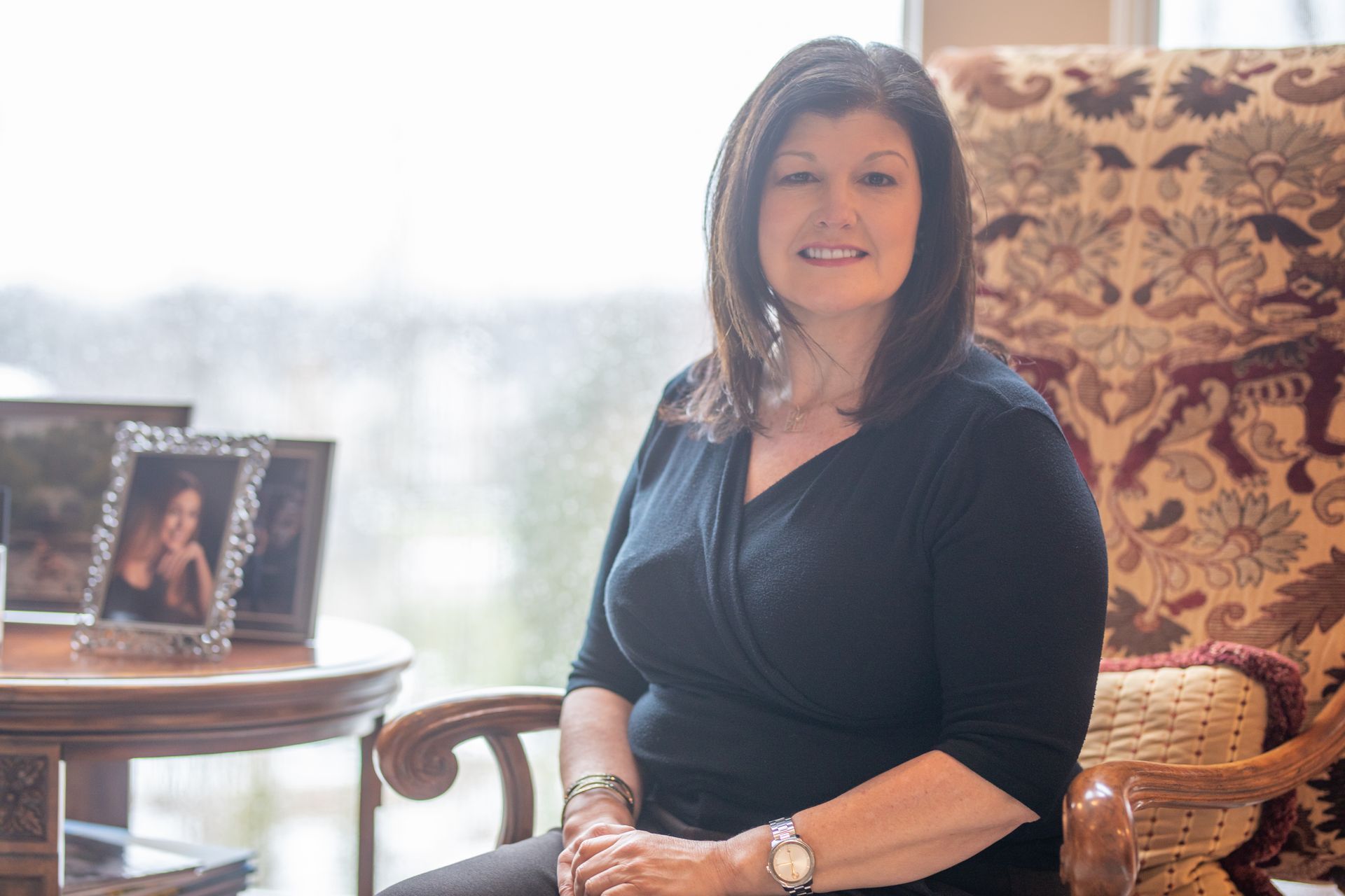 A woman in a black shirt sits in a patterned armchair next to a small table with framed photos by a bright window.