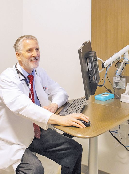 A smiling professional in a white coat sits at a desk using a computer mouse in a clinical office setting.