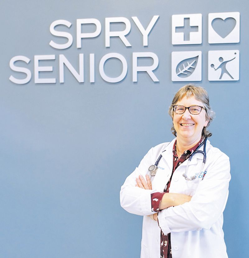 A smiling medical professional in a white coat and stethoscope stands before a wall sign reading 