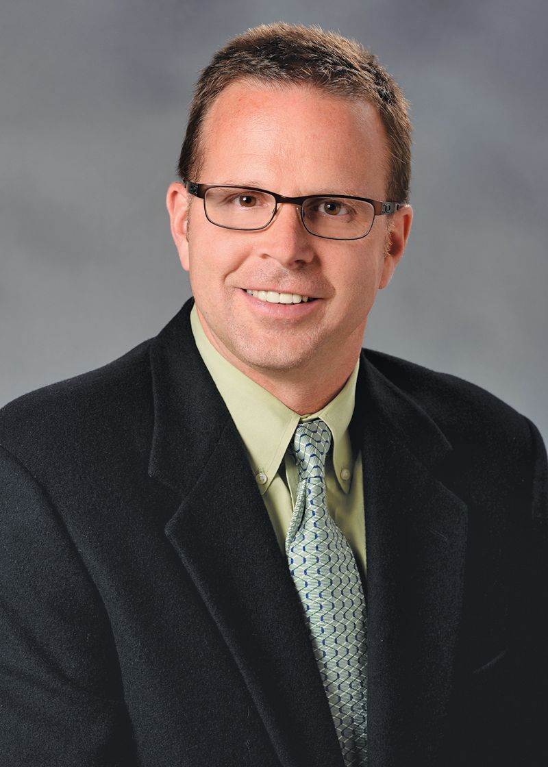 Headshot of a smiling person wearing glasses, a light green shirt, a patterned tie, and a dark blazer against a grey wall.