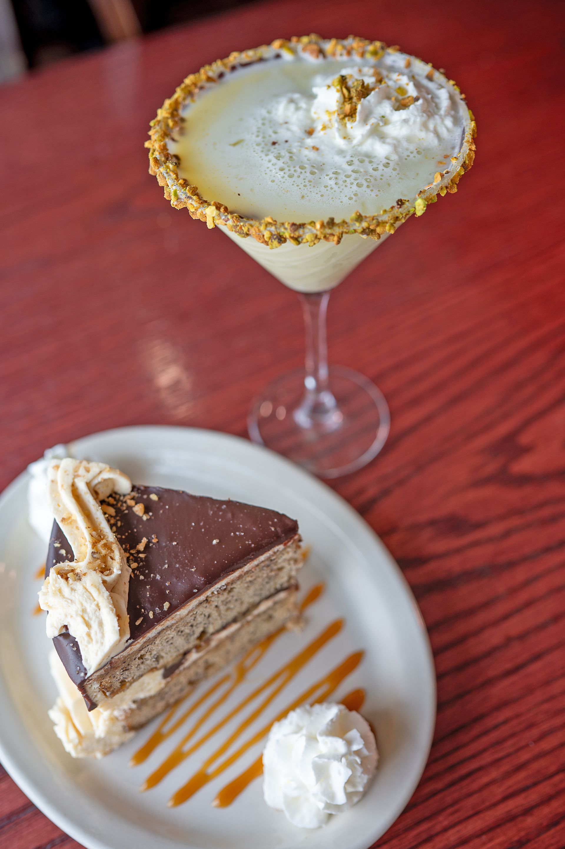 A slice of chocolate-frosted layer cake with whipped cream next to a rimmed cocktail in a martini glass on a red table.
