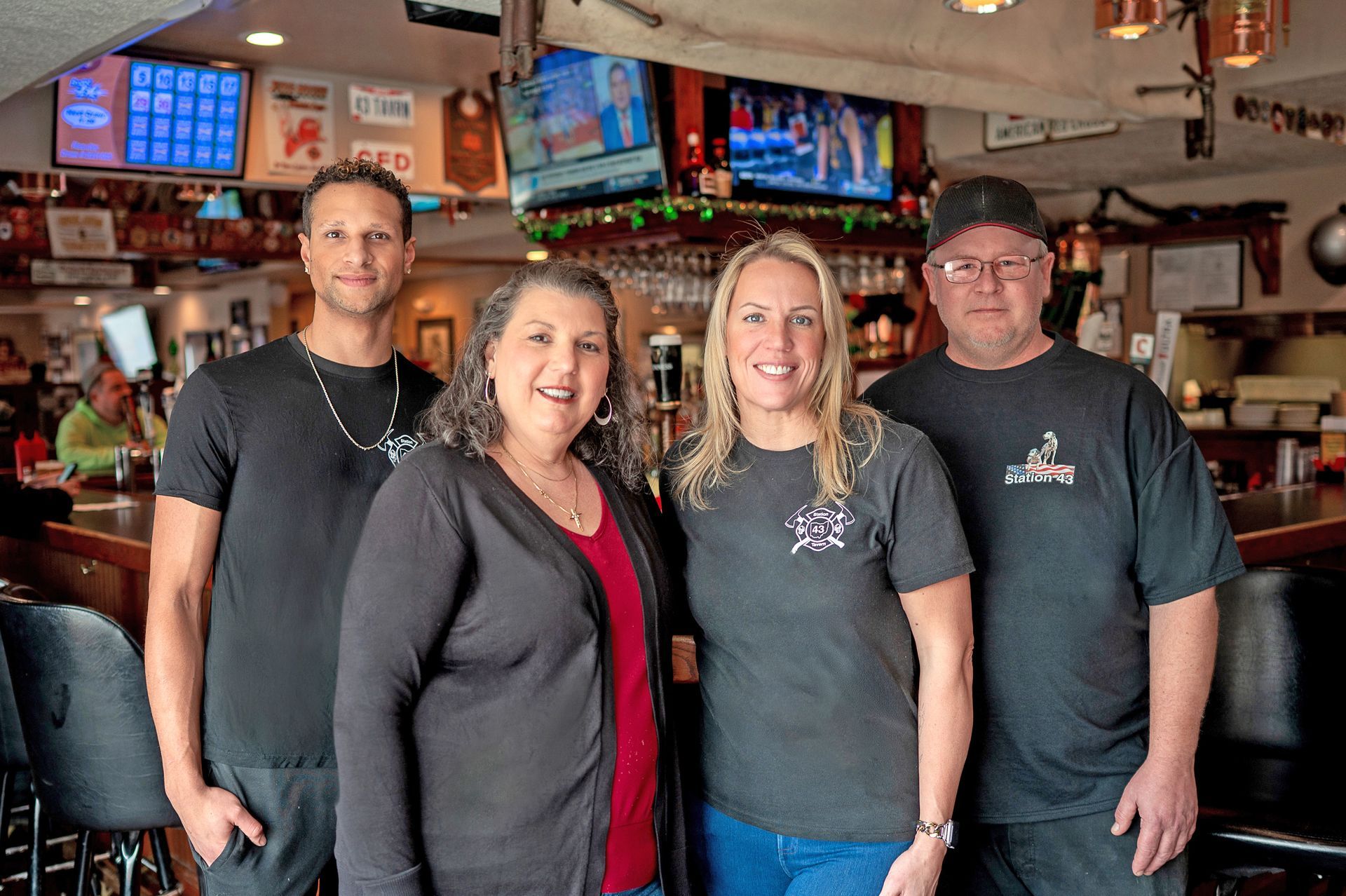 Four people pose together in a bar setting, smiling toward the camera behind a wooden counter.