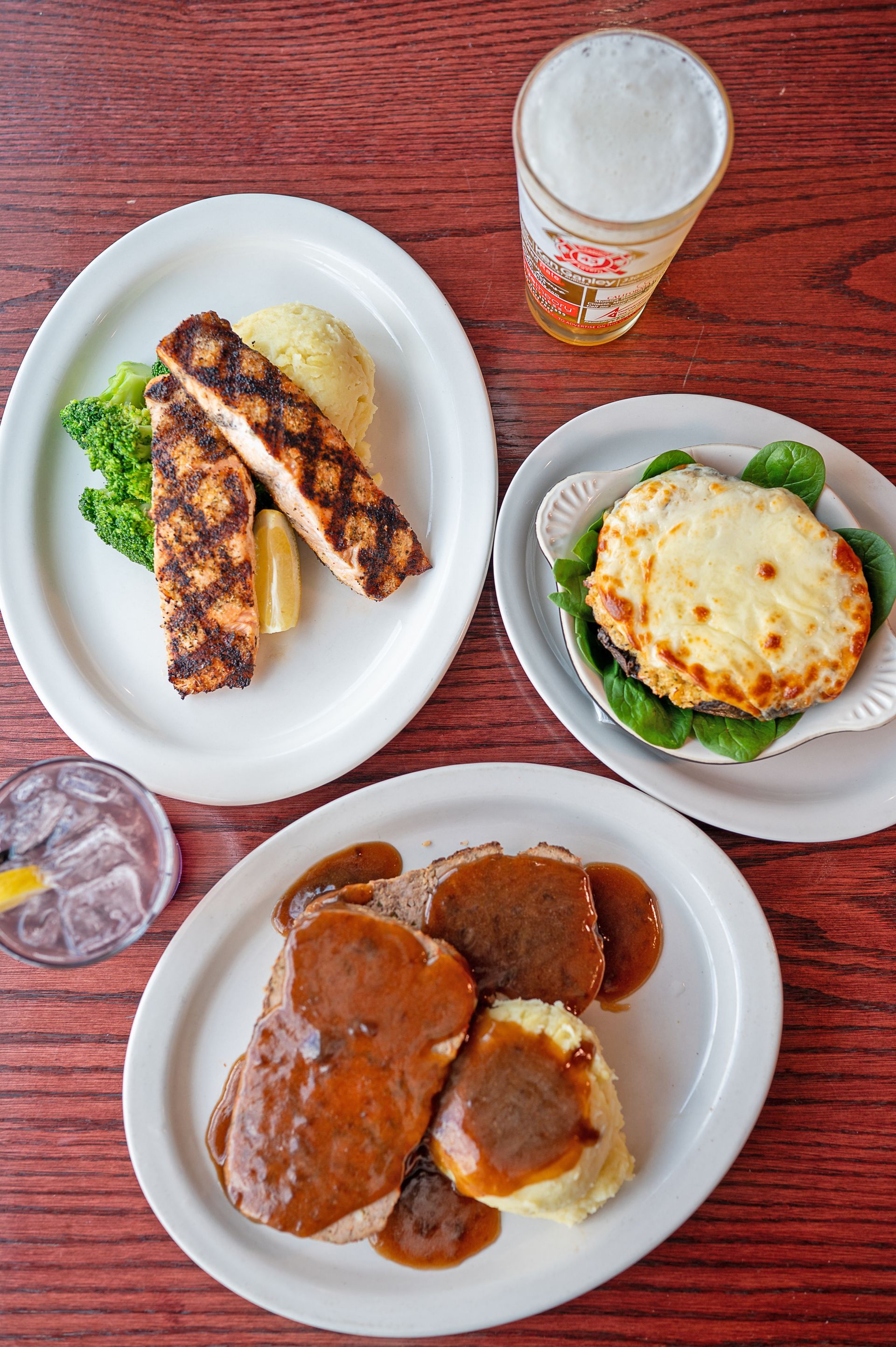 Three plates of food, including grilled fish, a baked dish, and meat with gravy, alongside a glass of beer and iced water.