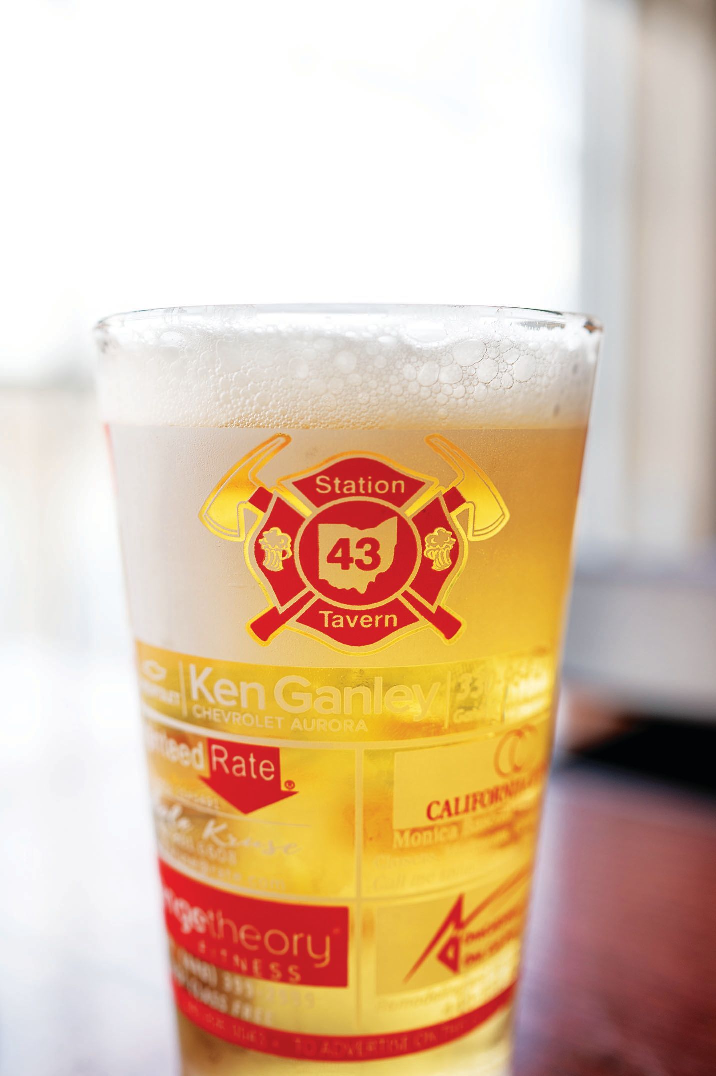 A clear glass of beer with a red fire-department-style logo featuring the number 43 and various corporate advertisements.