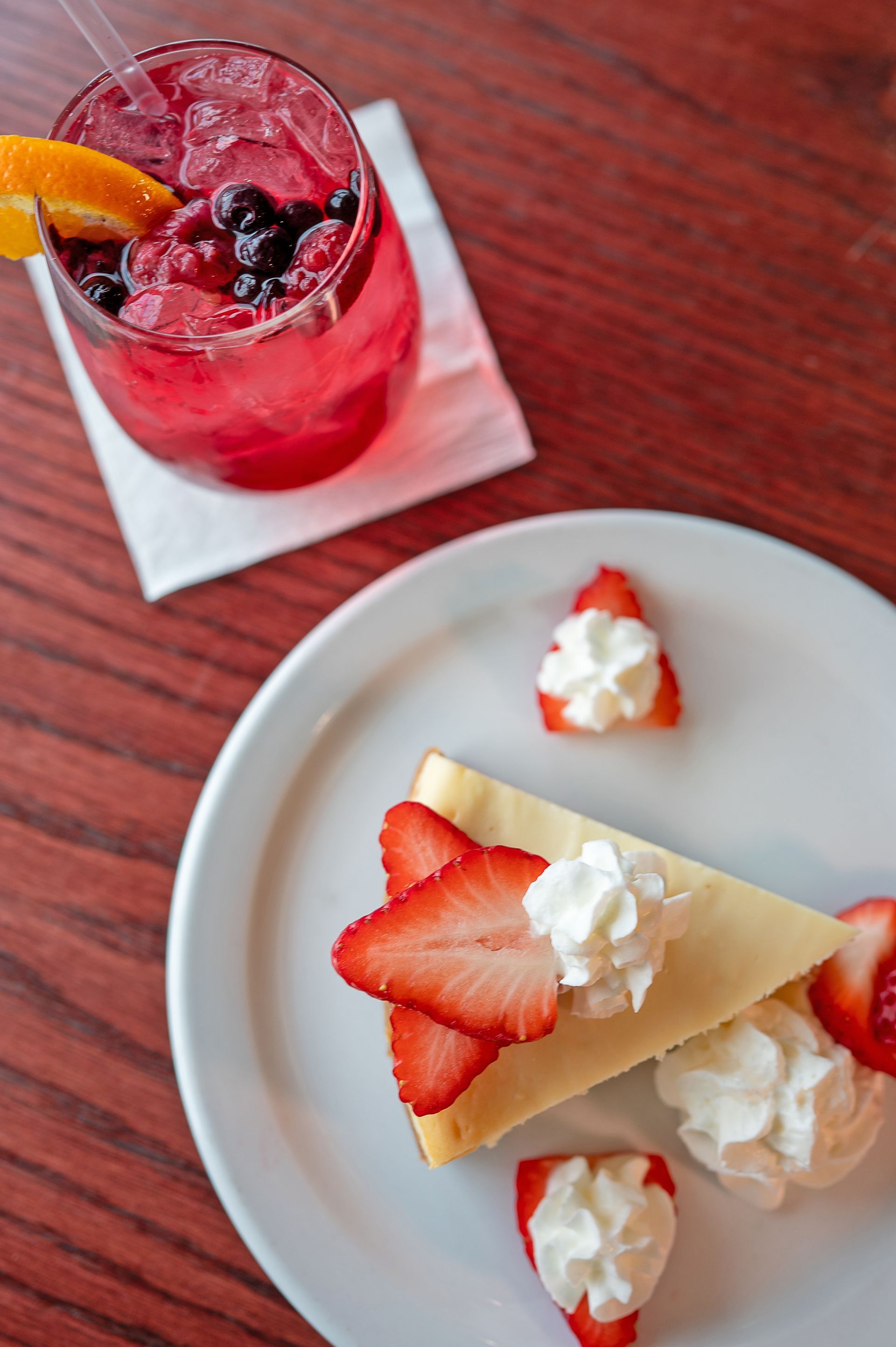 A slice of cheesecake topped with strawberries and whipped cream, next to a red berry drink on a wooden table.