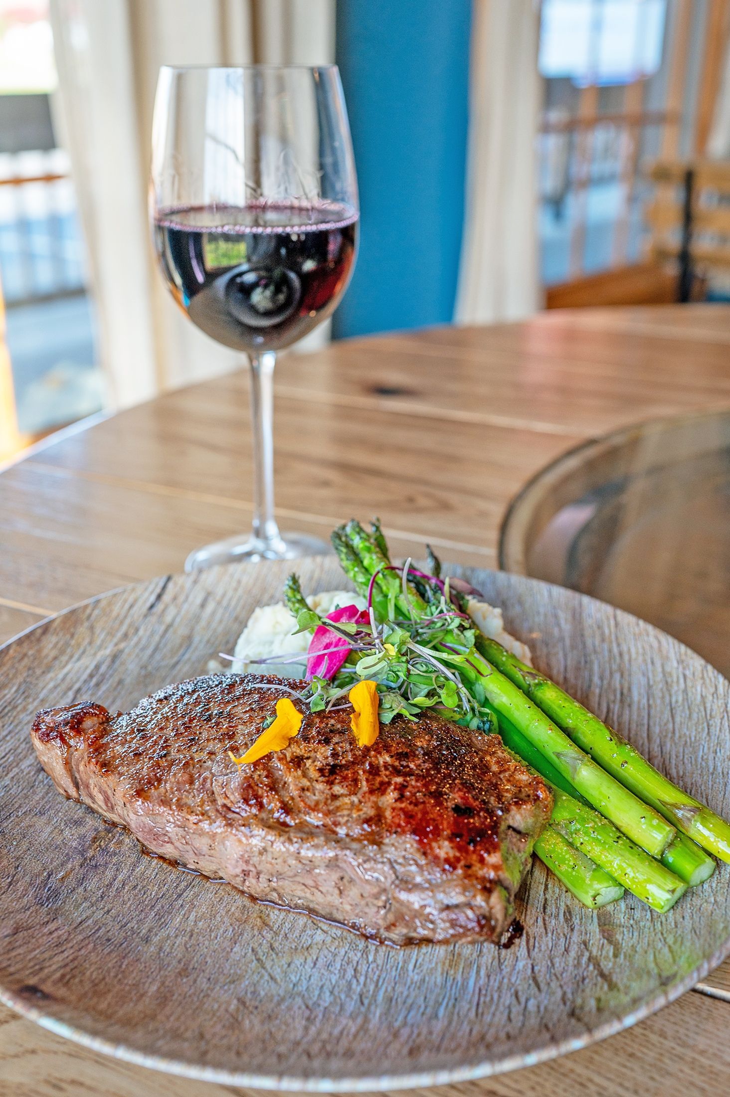 A grilled steak served on a wooden plate with asparagus and mashed potatoes, accompanied by a glass of red wine.