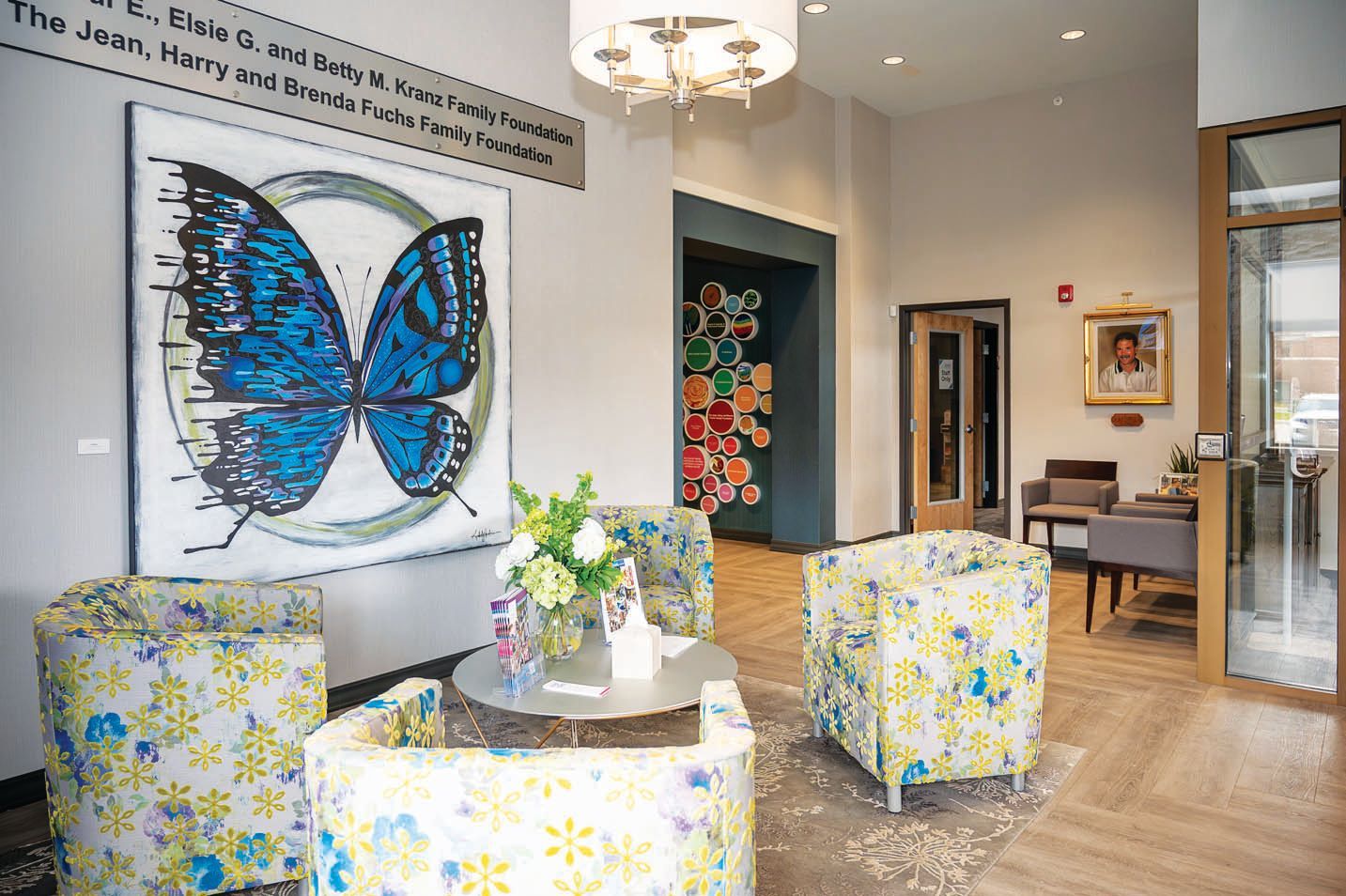 A bright, modern lobby features three floral armchairs, a central side table with flowers, and a large blue butterfly art.
