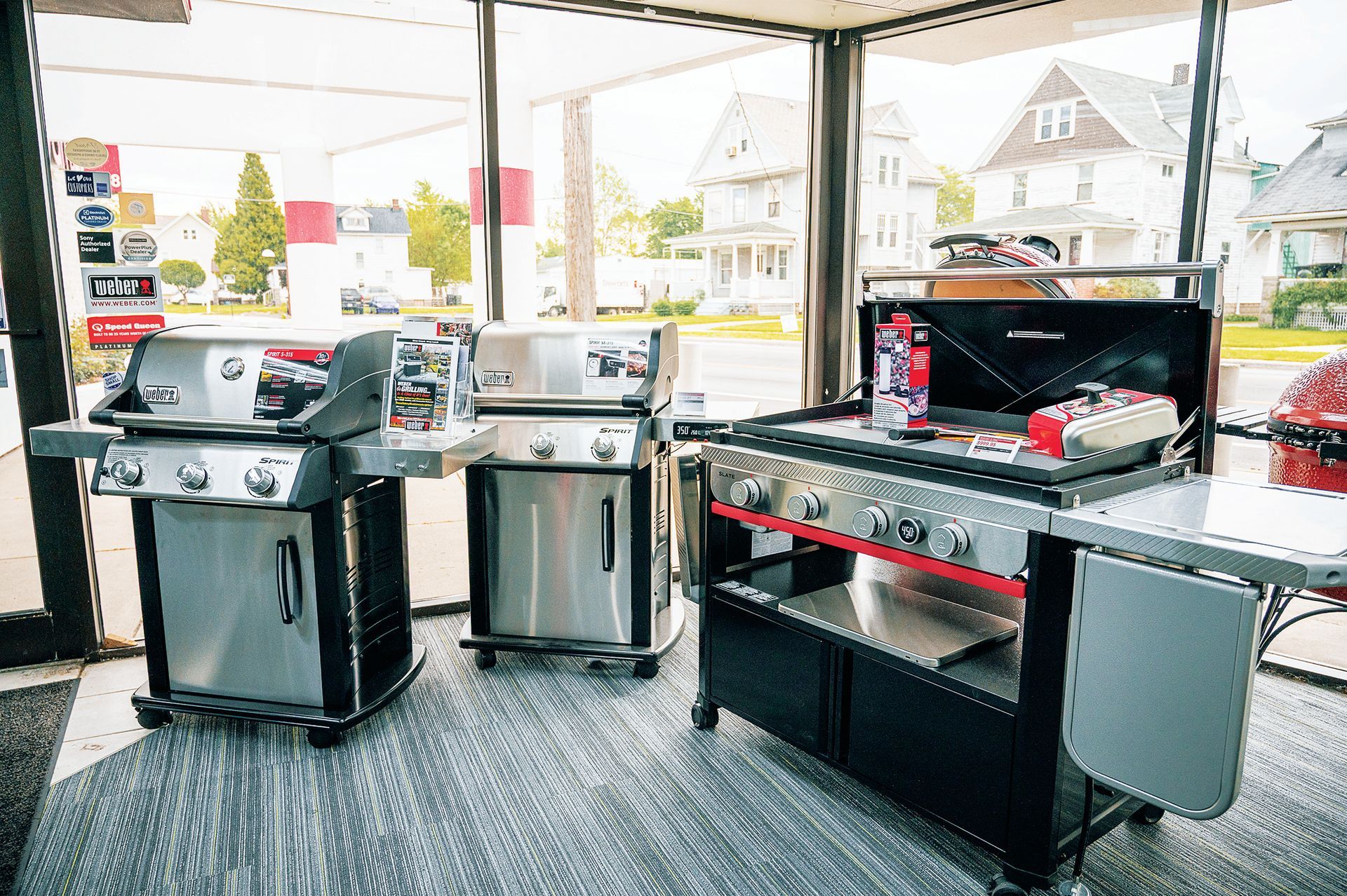 Three stainless steel outdoor grills arranged in a row inside a showroom with houses visible in the background.