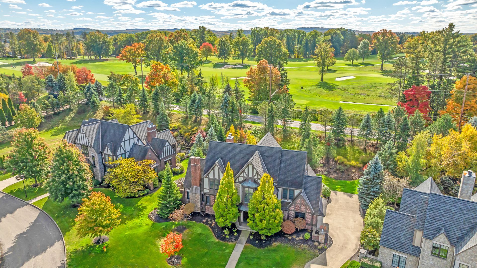 Aerial view of upscale homes with dark roofs surrounded by autumn trees and a golf course under a blue, cloudy sky.