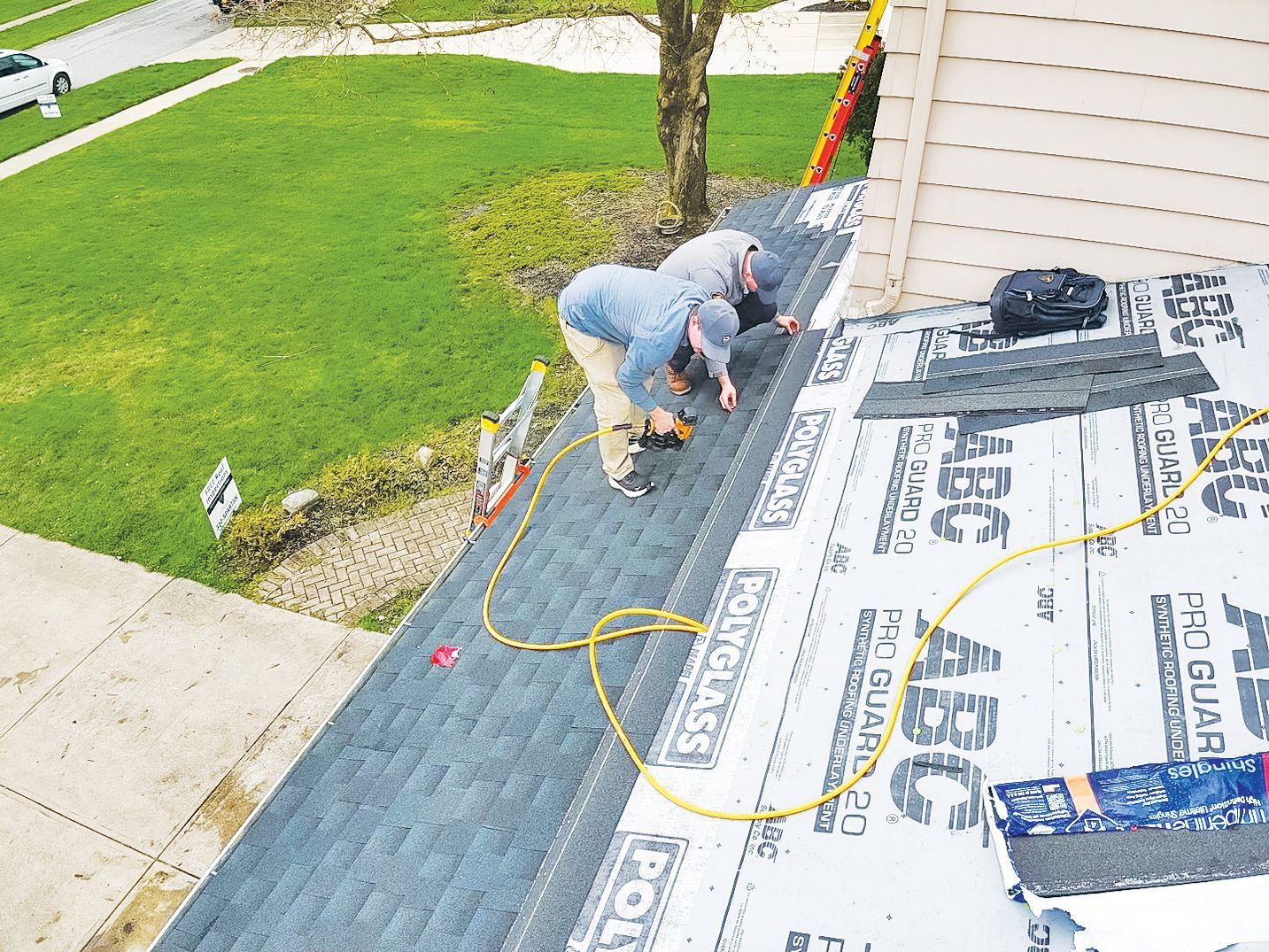 Two construction workers install dark roof shingles over white underlayment on a residential roof.